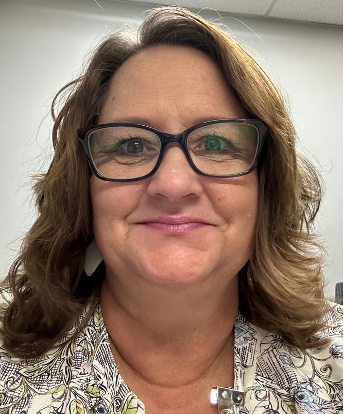 A woman with wavy, shoulder-length brown hair wearing glasses and a patterned blouse, smiling at the camera indoors.
