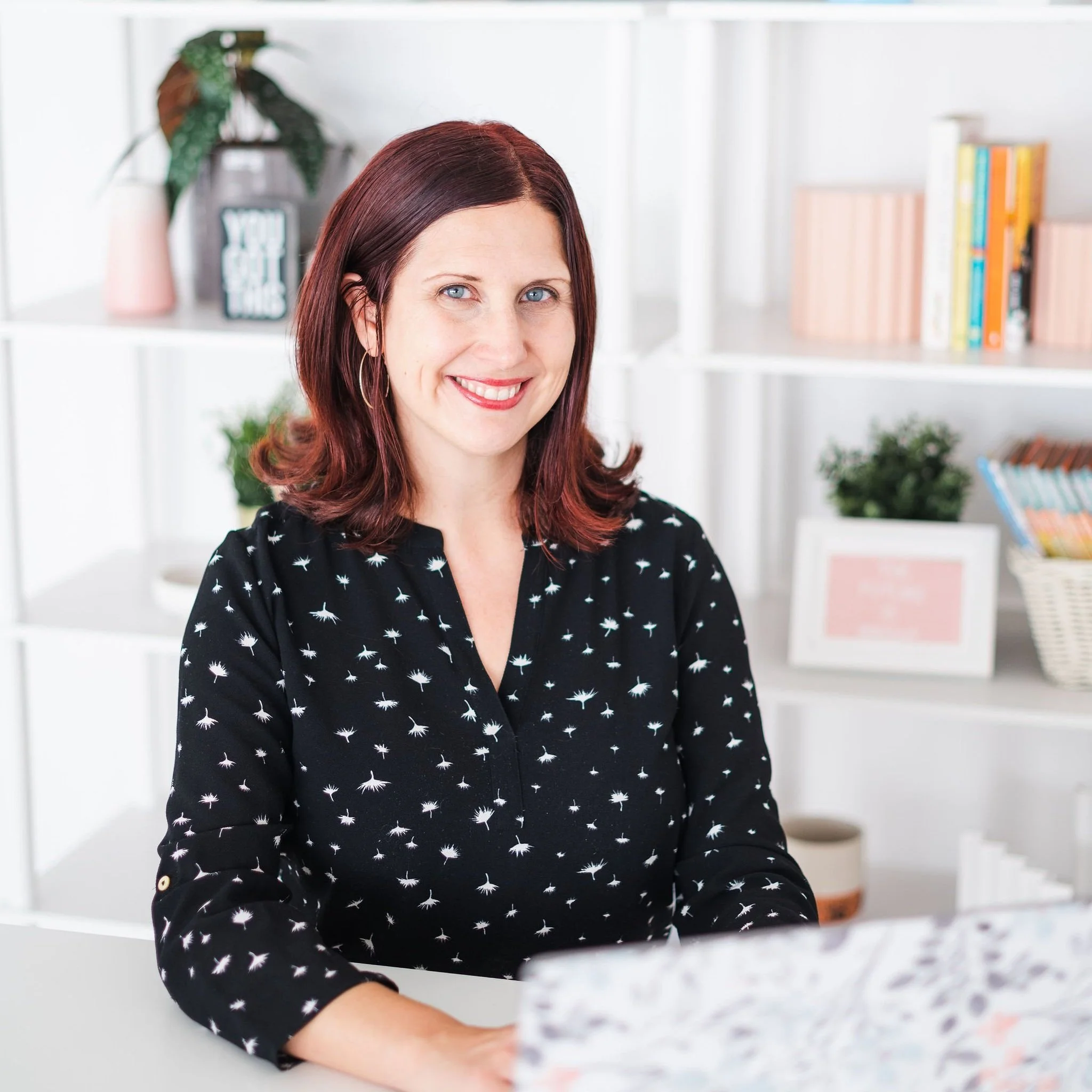 A woman with shoulder-length red hair and blue eyes smiling at the camera, sitting at a white desk in a bright room with a white bookshelf and framed picture in the background.