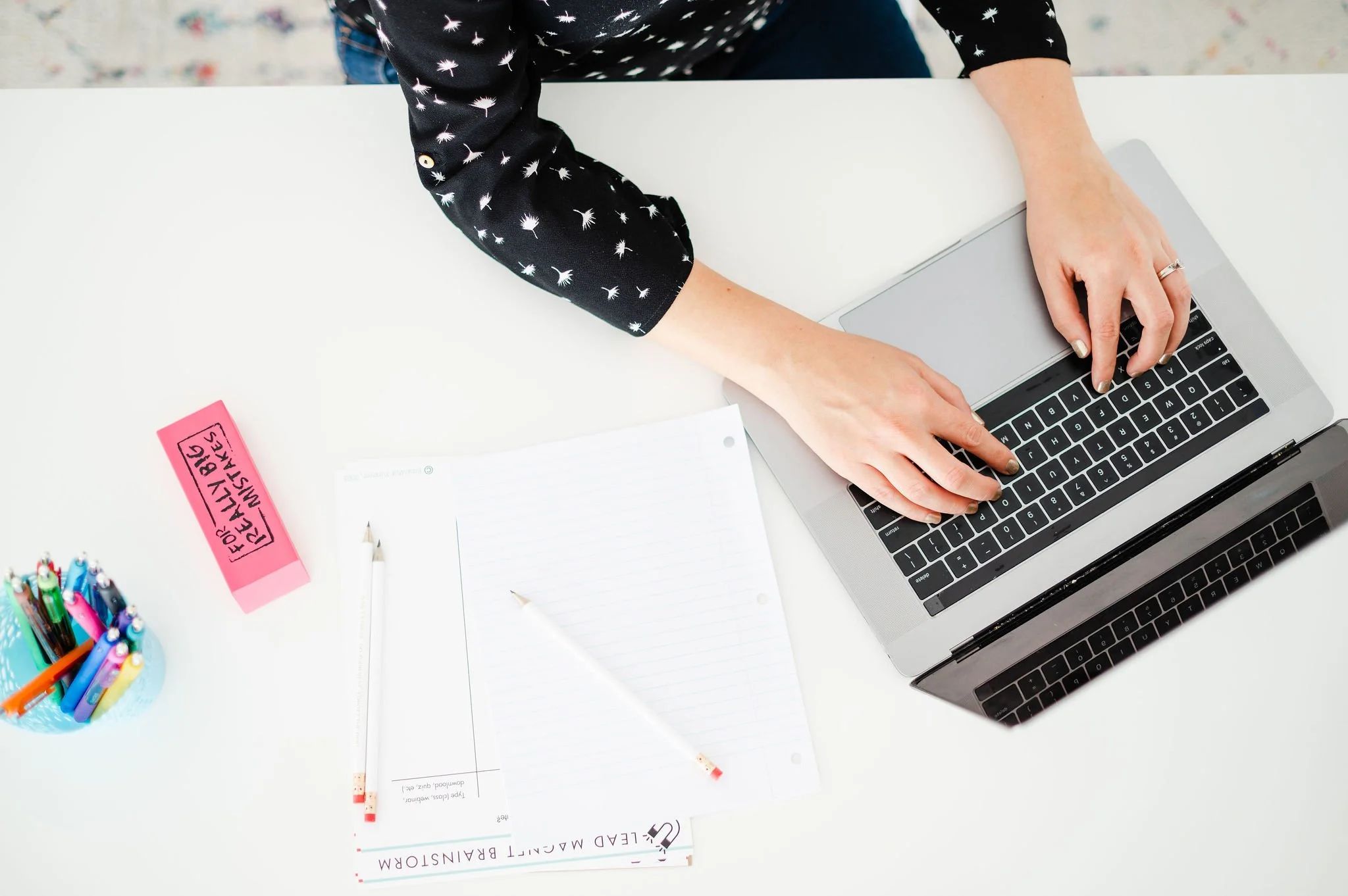 A person with a black sweater with white dandelion patterns typing on a silver laptop on a white desk. On the desk are notebooks, a white pen, and a blue cup filled with colorful pens. A pink sticky note reads 'FOR ALL YOUR EMAIL & MESSAGING'.