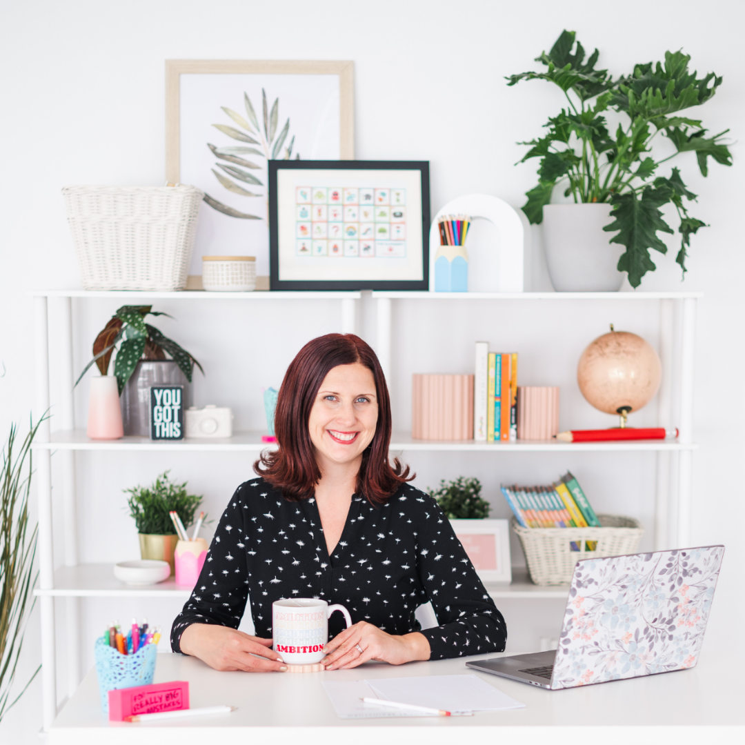 A woman sitting at a white desk in a colorful and decorated home office, smiling and holding a mug. Behind her is a white bookshelf with various books, plants, art, and decorative items.