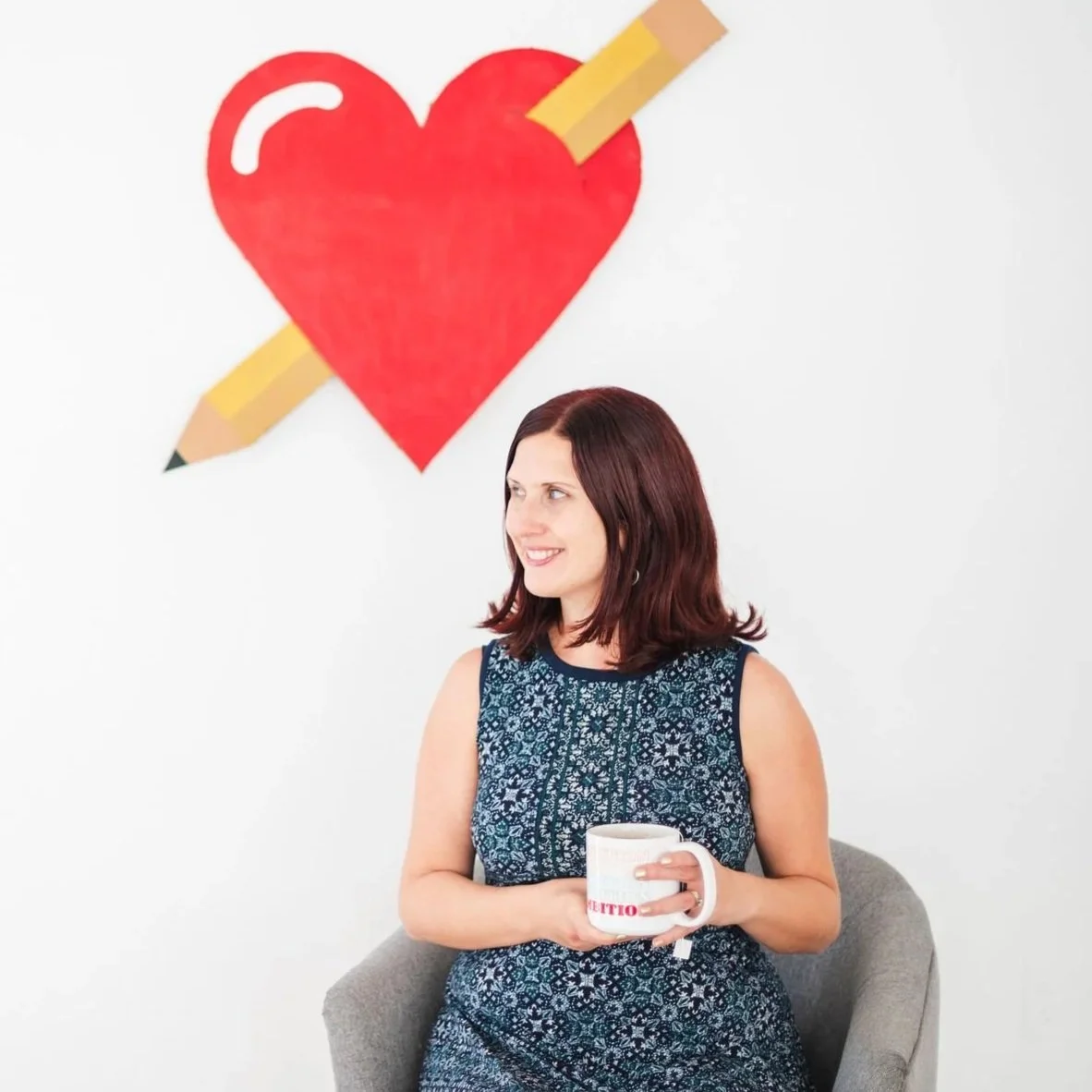 A woman with dark red hair, wearing a sleeveless blue patterned dress, sitting on a gray chair, holding a white mug, smiling and looking to the right, with a white wall and a large red heart with a yellow pencil behind it painted on the wall.