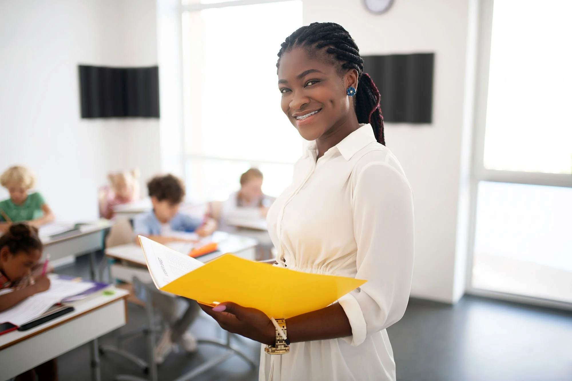 A smiling woman with dark skin, wearing a white blouse and blue earrings, standing in a classroom holding a yellow folder, with children working at desks in the background.