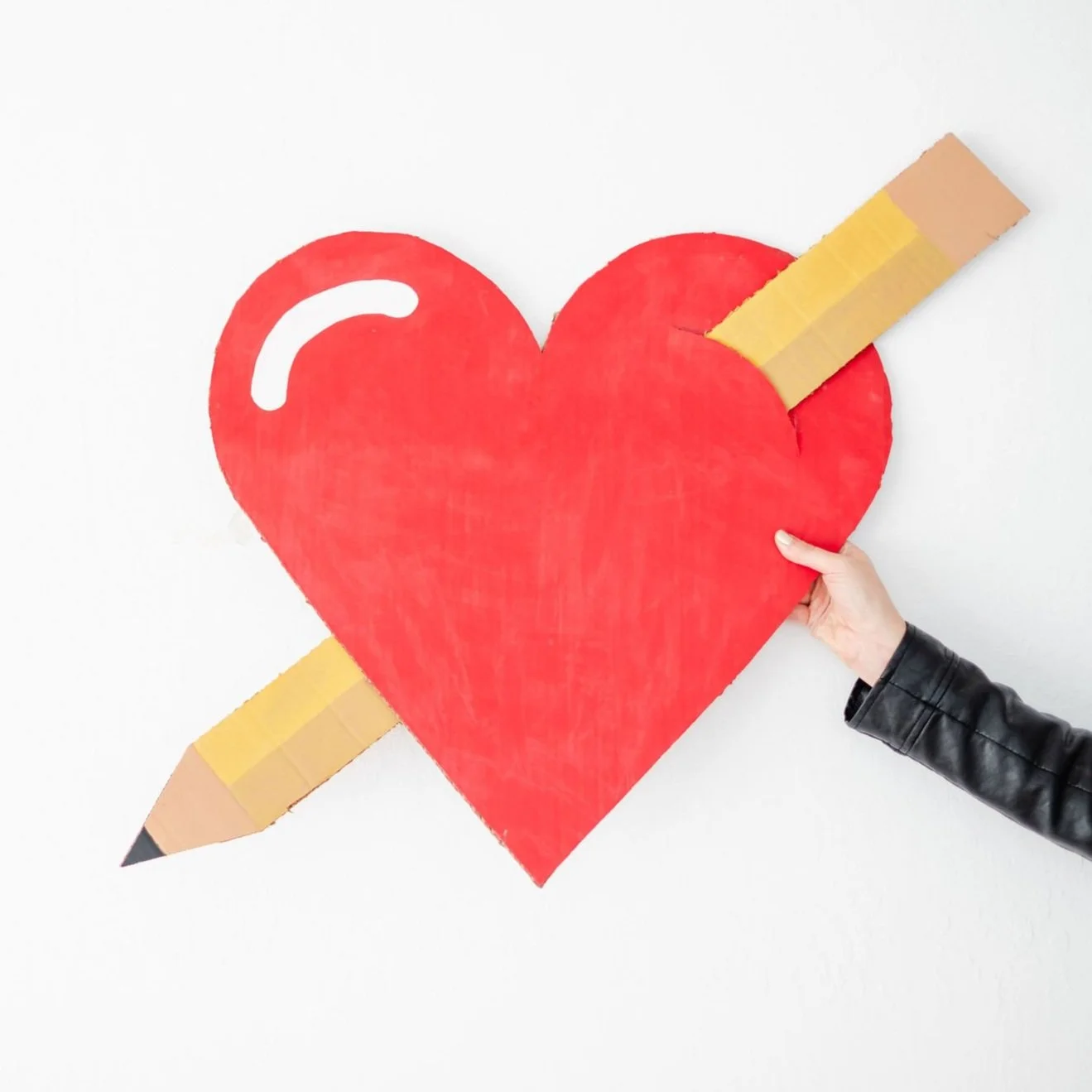 Hand holding a large red heart with a pencil on it, representing a love for teaching.