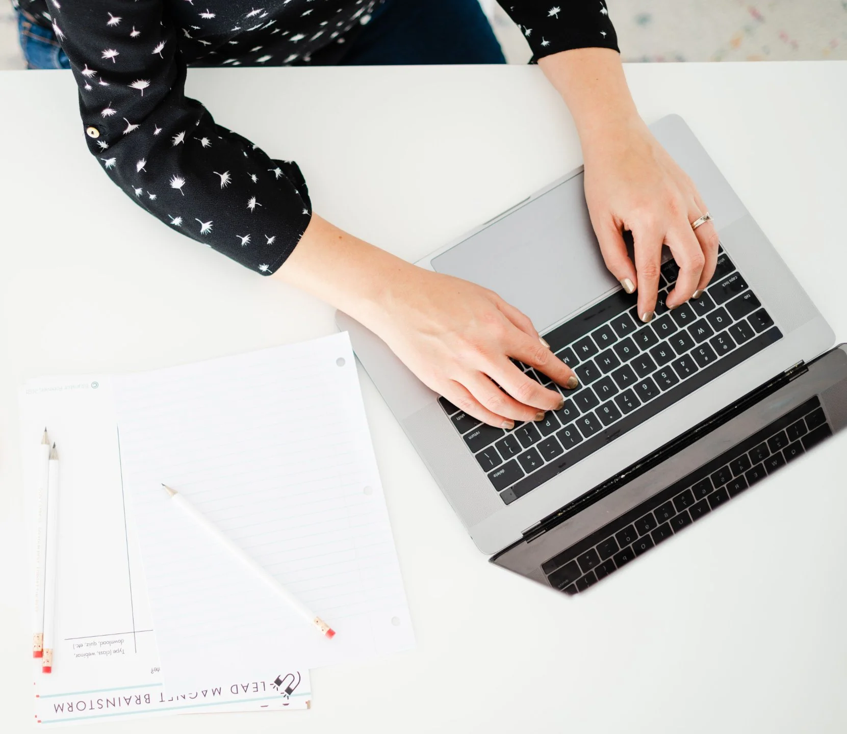 A person wearing a black shirt with white dandelion patterns working on a laptop at a white desk. There are lined notepads, a white pen with red accents, and additional sheets of paper on the desk.