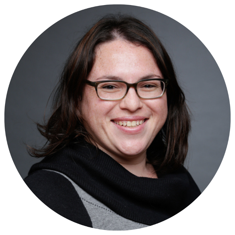 A woman with shoulder-length dark hair, wearing glasses and a black sweater, smiling against a gray background.