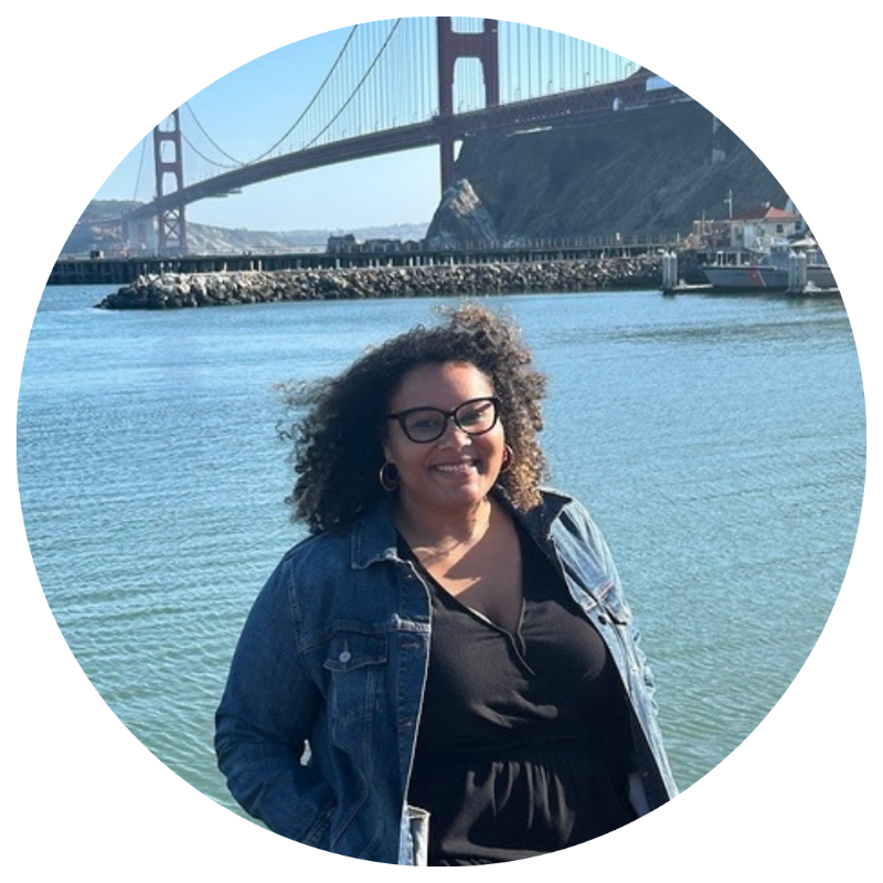 Woman with curly hair and glasses smiling near water with the Golden Gate Bridge in the background.