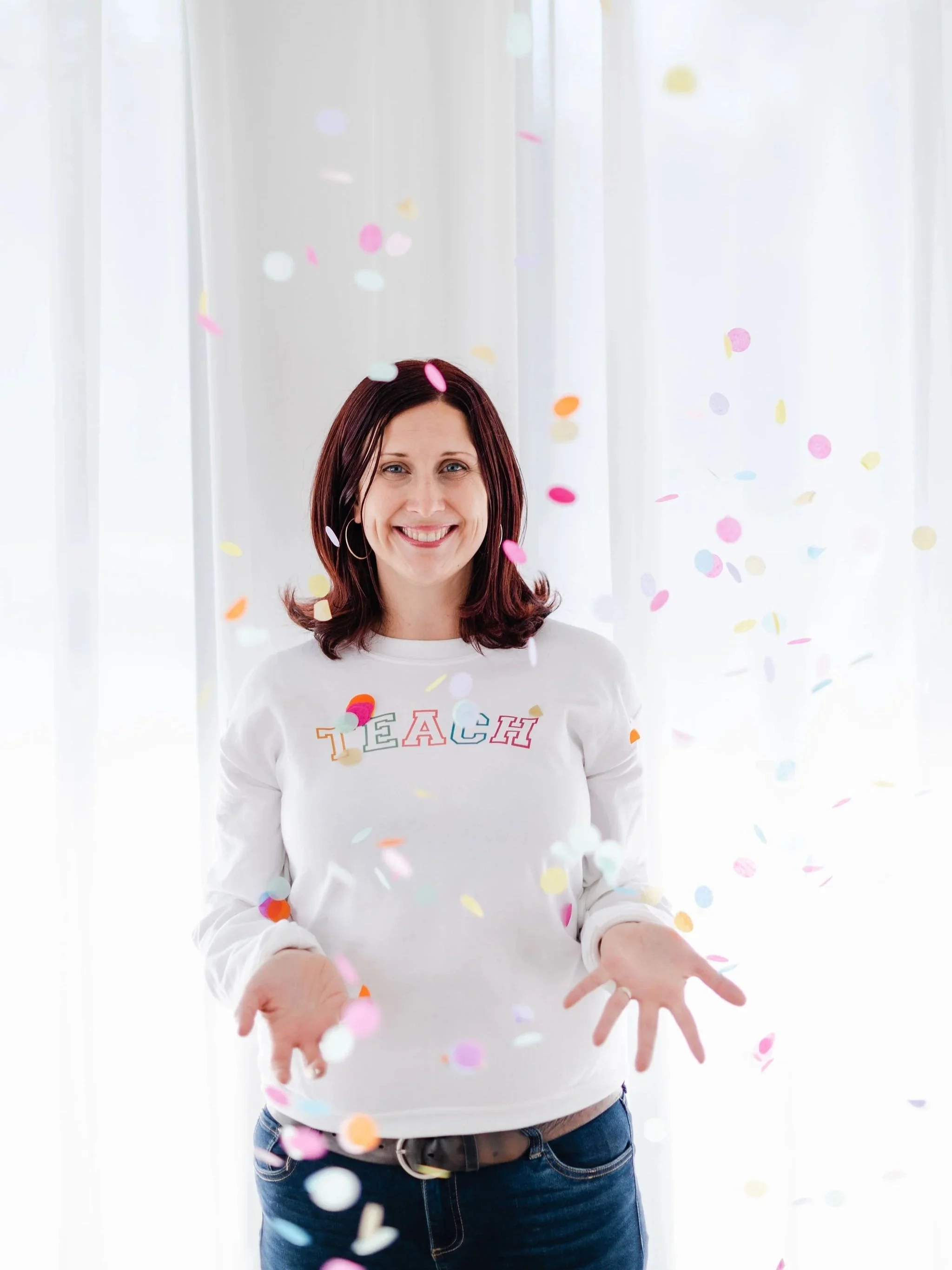 Woman with brown hair wearing a white sweatshirt with the word 'TEACH' in colorful letters, standing in front of white curtains, surrounded by falling colorful confetti, and smiling.
