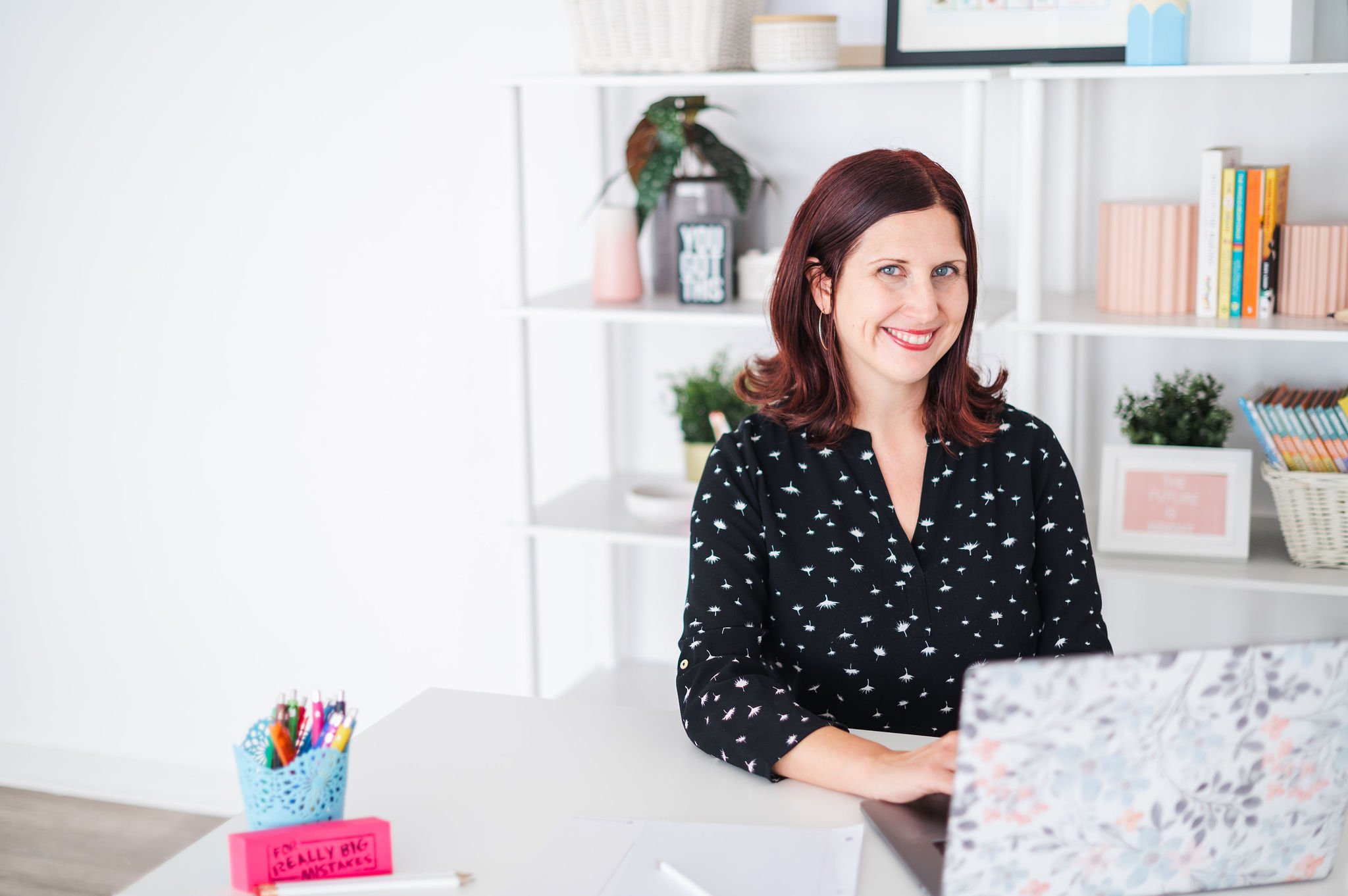A woman with shoulder-length red hair, wearing a black blouse with a white dandelion pattern, sitting at a white desk with a floral notebook open and a silver pen nearby. In the background, there are white shelves with books, plants, and decorative items, and a white basket filled with colorful pens on the desk.