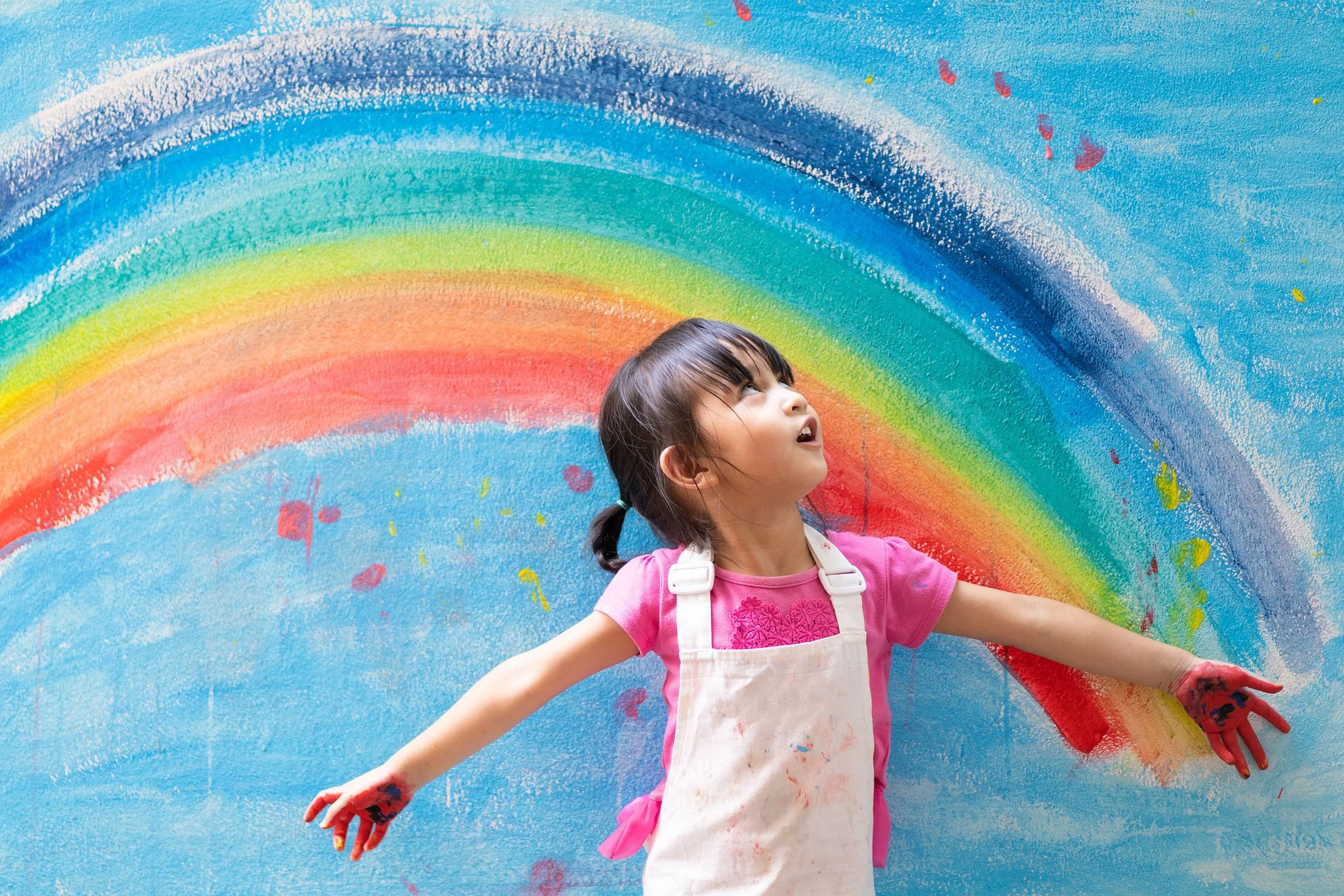 Young girl with brown hair wearing a pink shirt and white apron, standing in front of a colorful rainbow mural on a blue background and looking upward with arms outstretched.