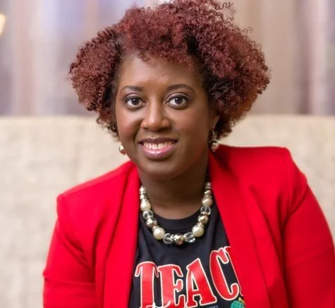 Portrait of a woman with curly red hair, wearing a red blazer, a black T-shirt with red and white text, and a pearl necklace, sitting indoors.