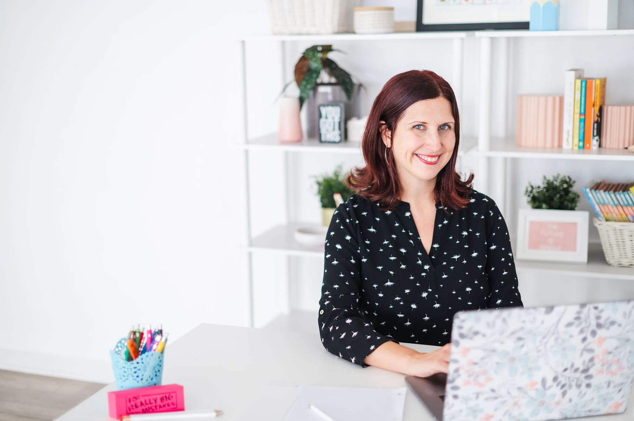 A woman with shoulder-length brunette hair smiling while sitting at a white desk with an open laptop. Behind her are white shelves with books, plants, and decorative items in a bright room.
