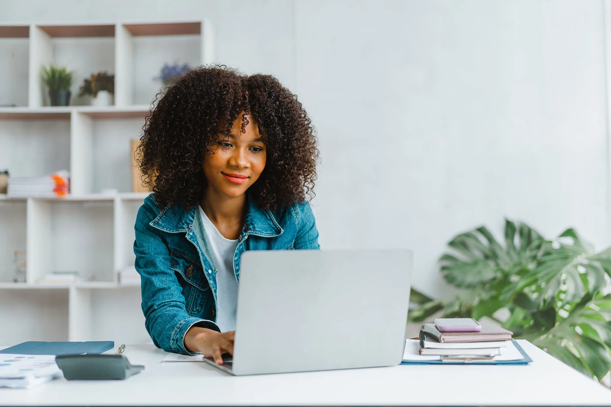 A woman with curly hair wearing a denim jacket working on a laptop at a white desk, with books and a potted plant nearby in a modern, minimalistic office space.