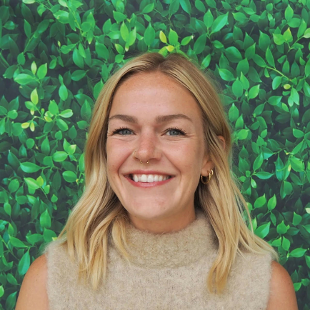 Smiling woman with shoulder-length blonde hair, nose ring, and gold hoop earrings, standing in front of a leafy green background.