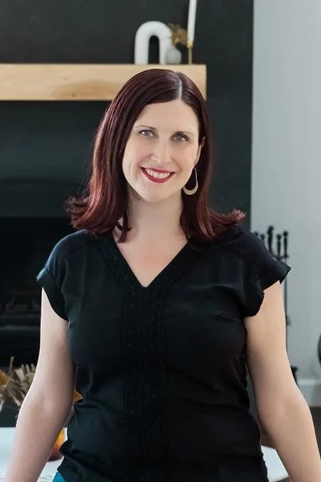 A woman with shoulder-length dark red hair, smiling, wearing a black top and hoop earrings, standing in a modern living room.