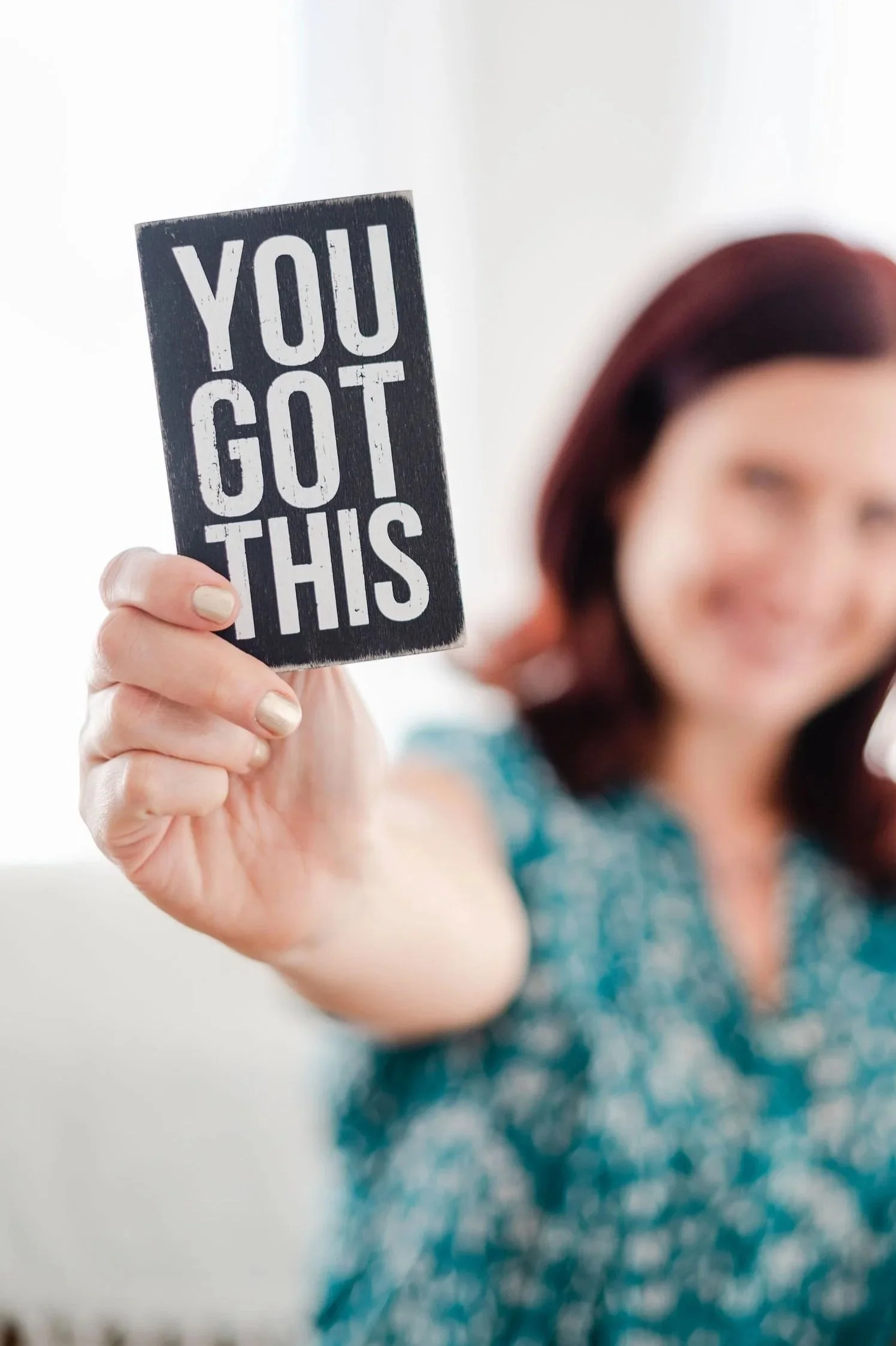 A woman with red hair holding a sign that says 'You Got This' towards the camera, with her face blurred in the background.