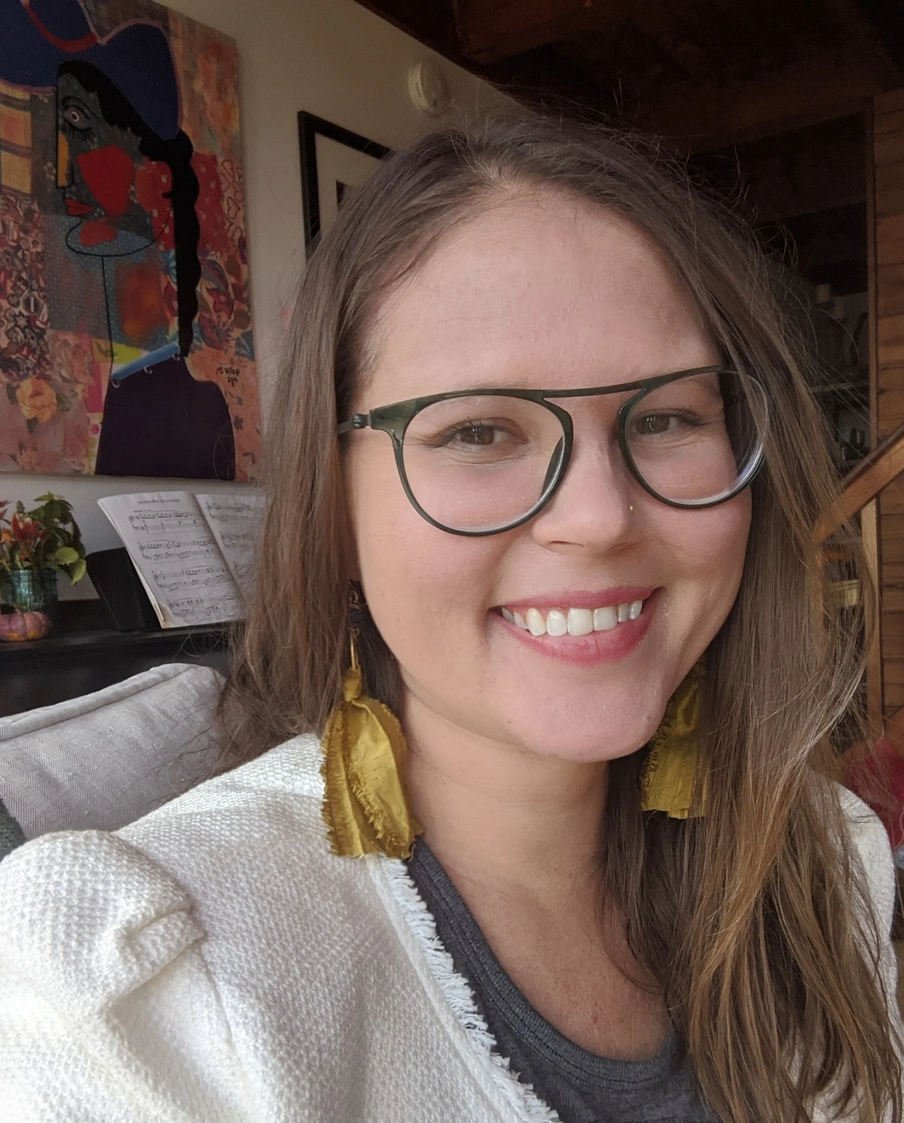 A smiling woman with long brown hair, wearing large round glasses, yellow earrings, a septum piercing, and a white blazer, indoors with a colorful painting and sheet music in the background.
