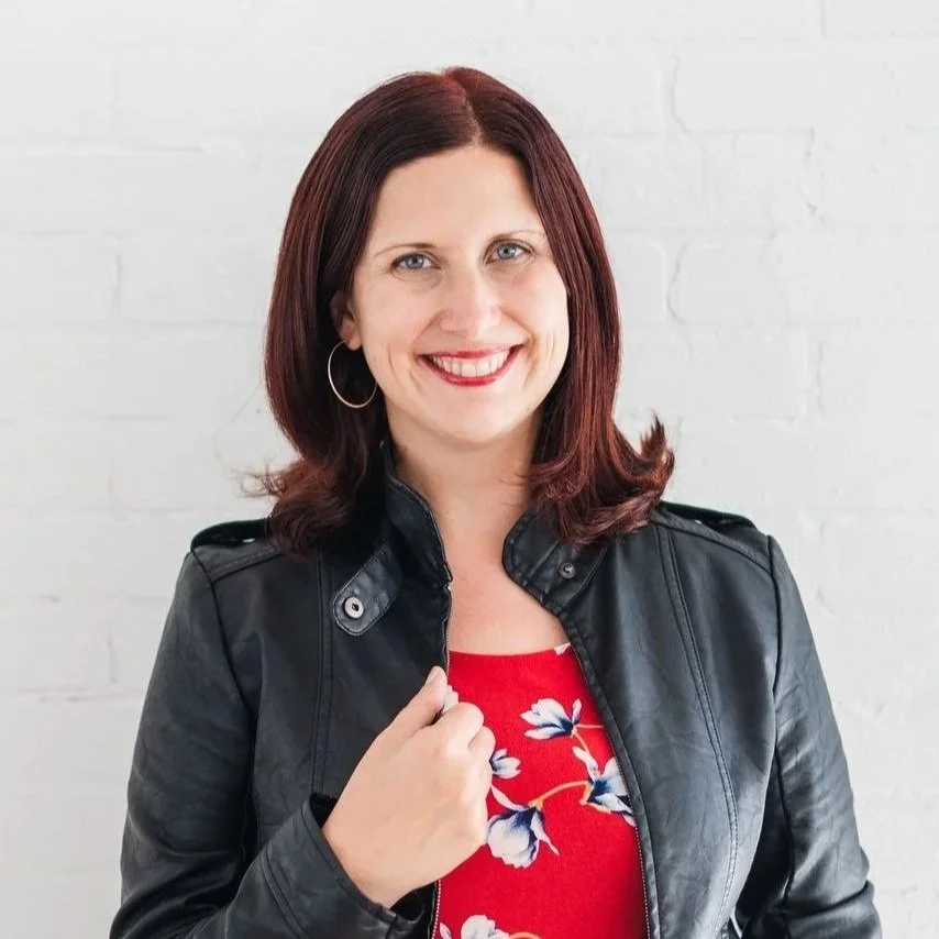 A woman with shoulder-length red hair smiling, wearing a black leather jacket over a red floral top, standing in front of a white brick wall.