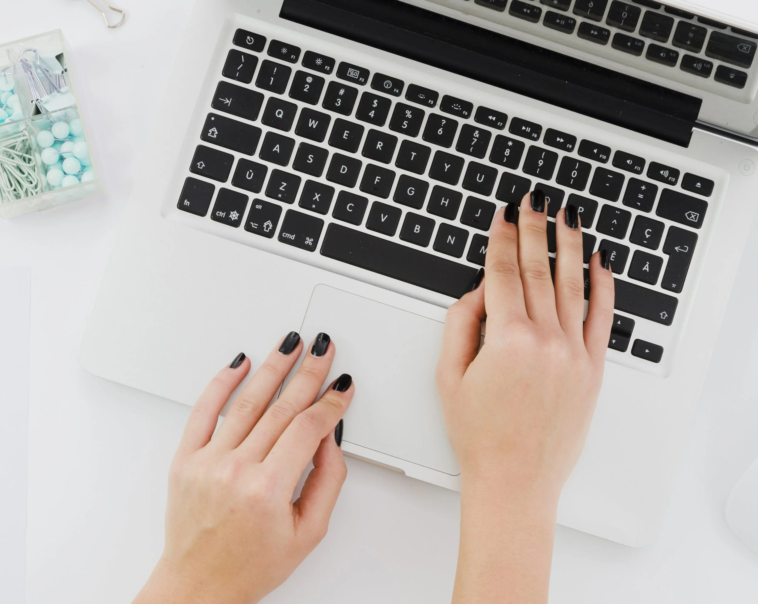 Person typing on a laptop keyboard with black painted nails, on a white desk with a small container of office supplies nearby.