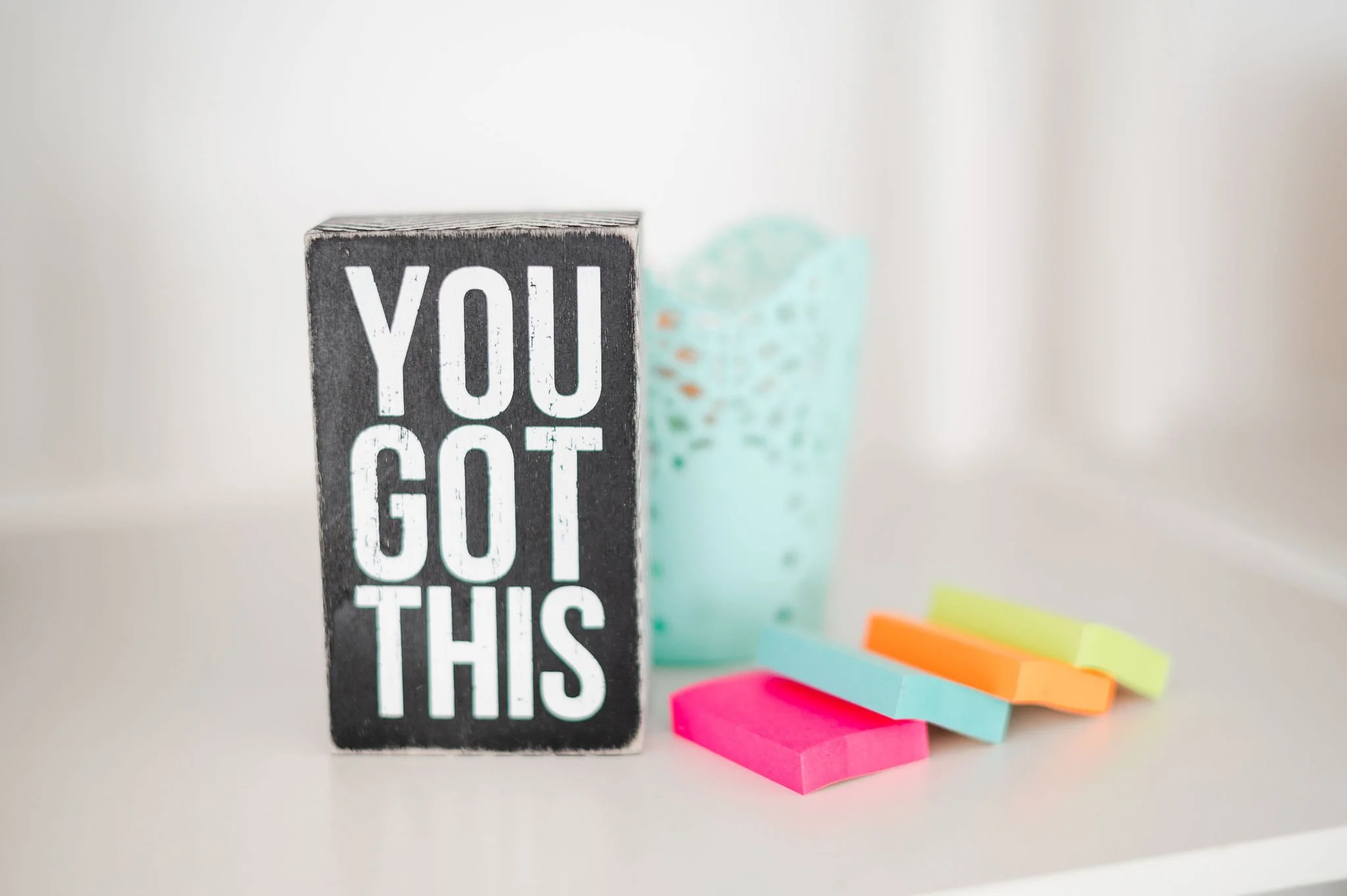 A black rectangular block with white bold text that says, 'YOU GOT THIS', positioned on a white surface. Behind it, there is a blurry container with blue-green dots and a few colorful sticky notes in pink, blue, orange, and yellow.