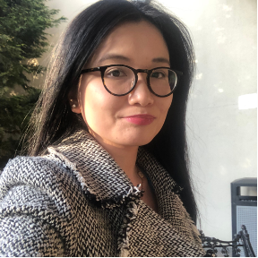 A woman with long black hair and glasses taking a selfie outdoors, wearing a checkered coat with a neutral background.