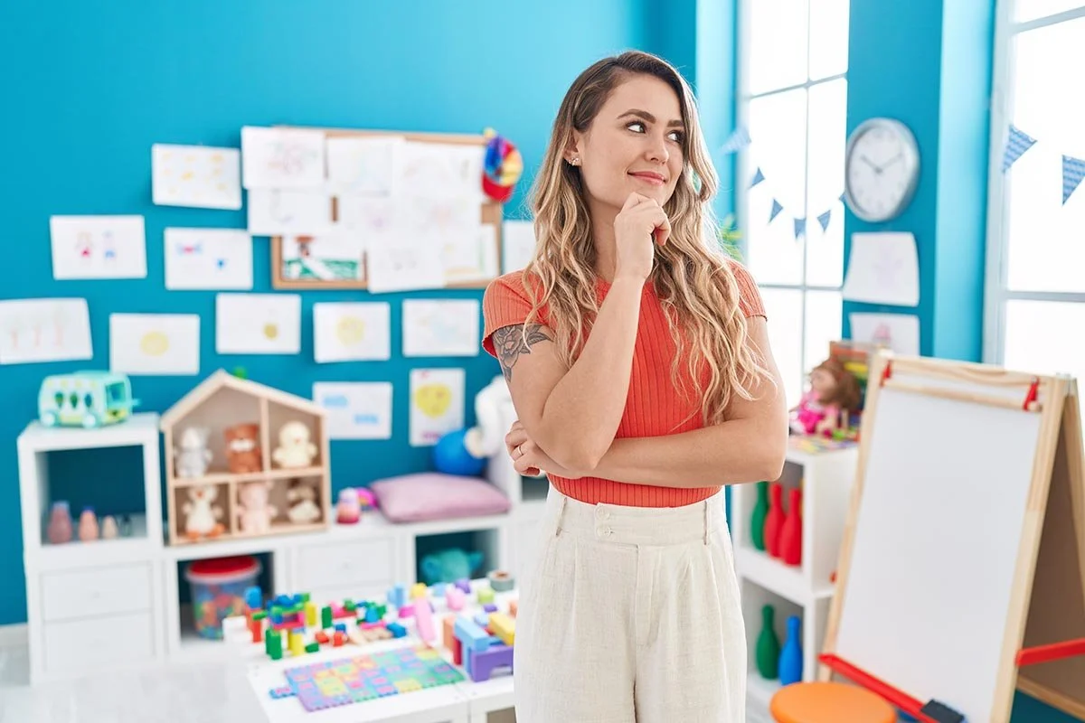 A woman teacher with long wavy hair and a tattoo on her arm stands in a colorful classroom, smiling thoughtfully with her hand on her chin. The classroom has bright blue walls, children's artwork, toys, and educational materials.