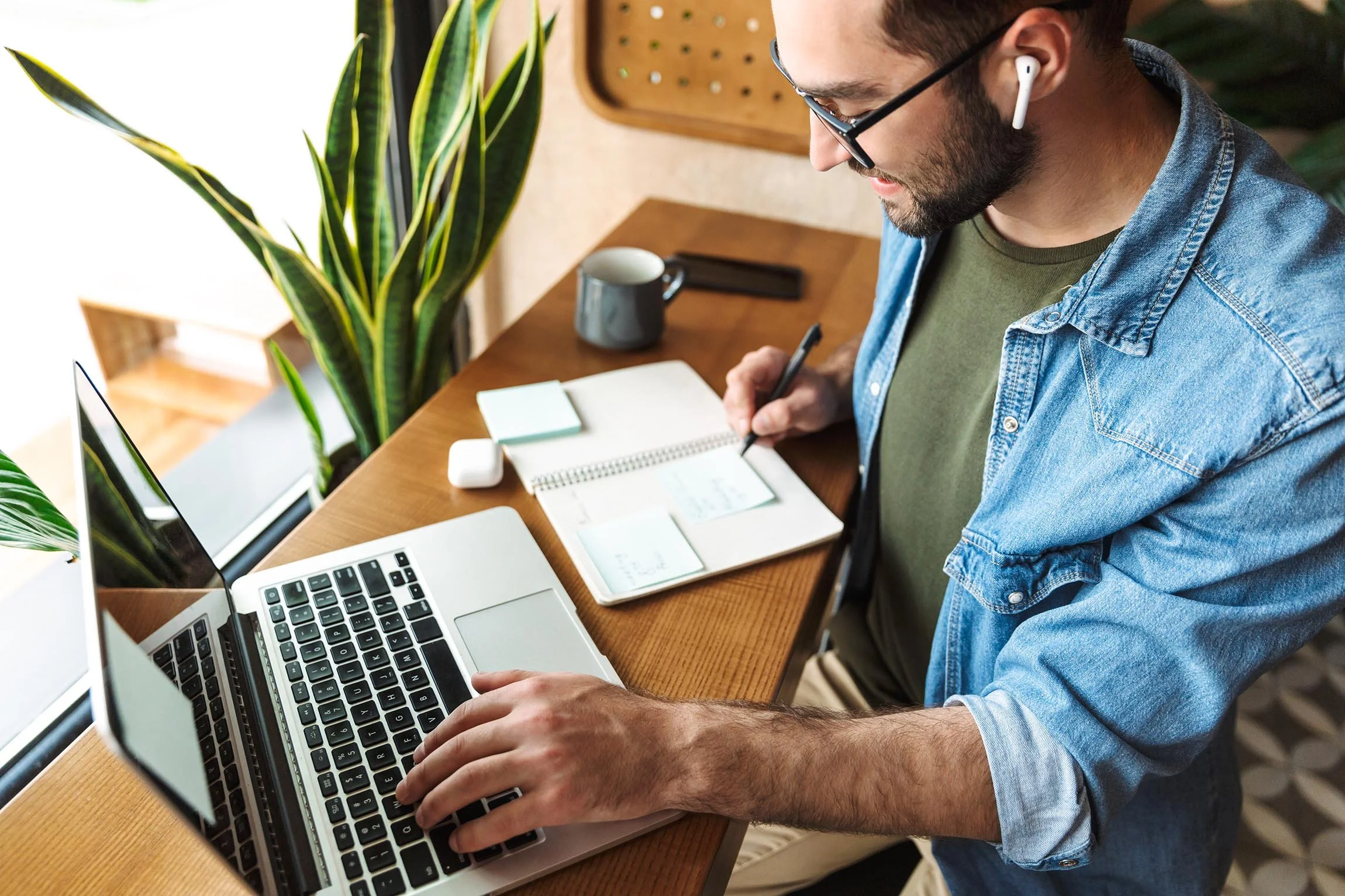 Young man working at a wooden desk with a laptop, notebook, pen, coffee mug, and plants near a window.