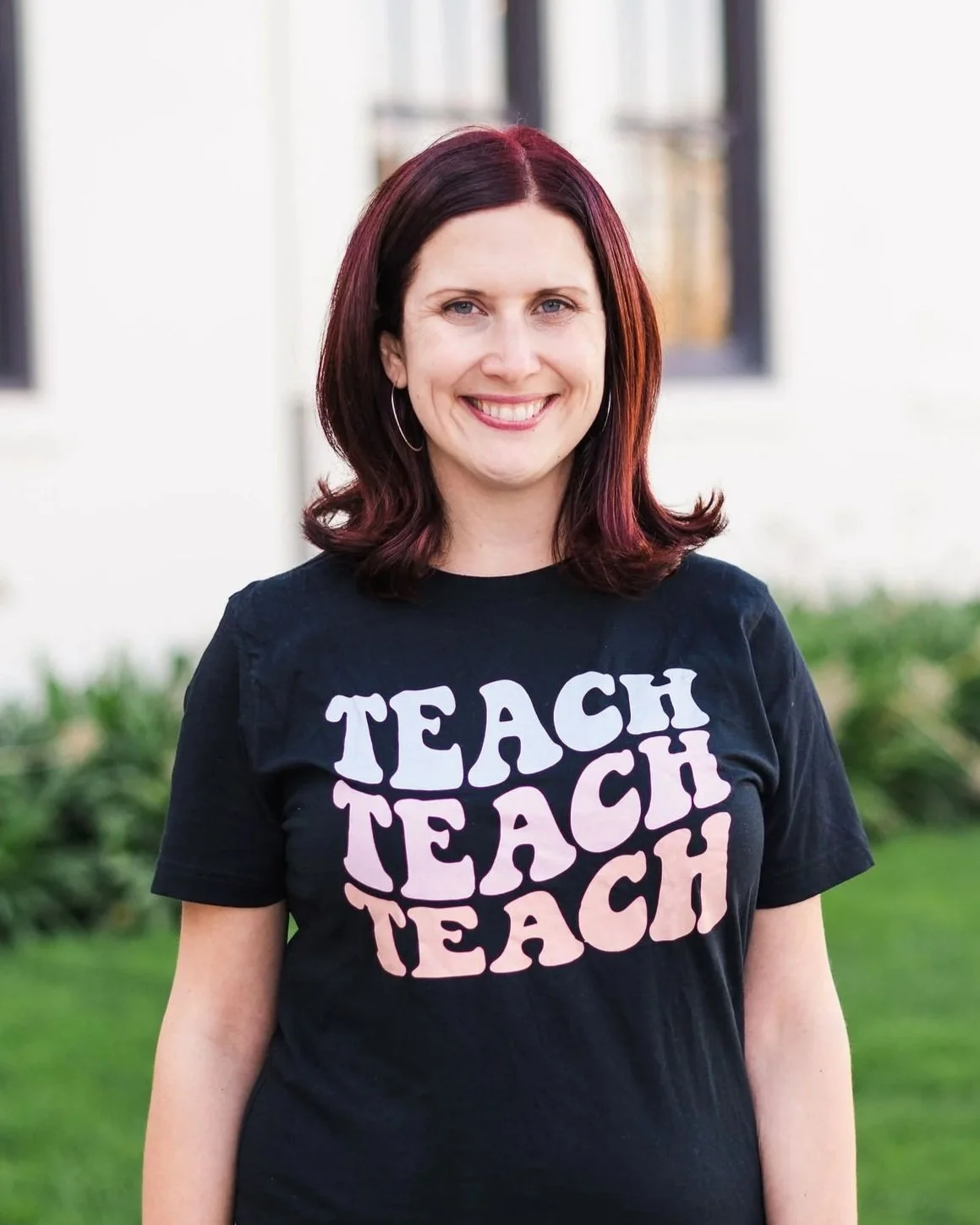 A woman with shoulder-length reddish-brown hair smiling, wearing a black T-shirt with the words 'TEACH' repeated four times in large white and pink letters, standing outdoors in front of a blurred white house with windows and greenery.