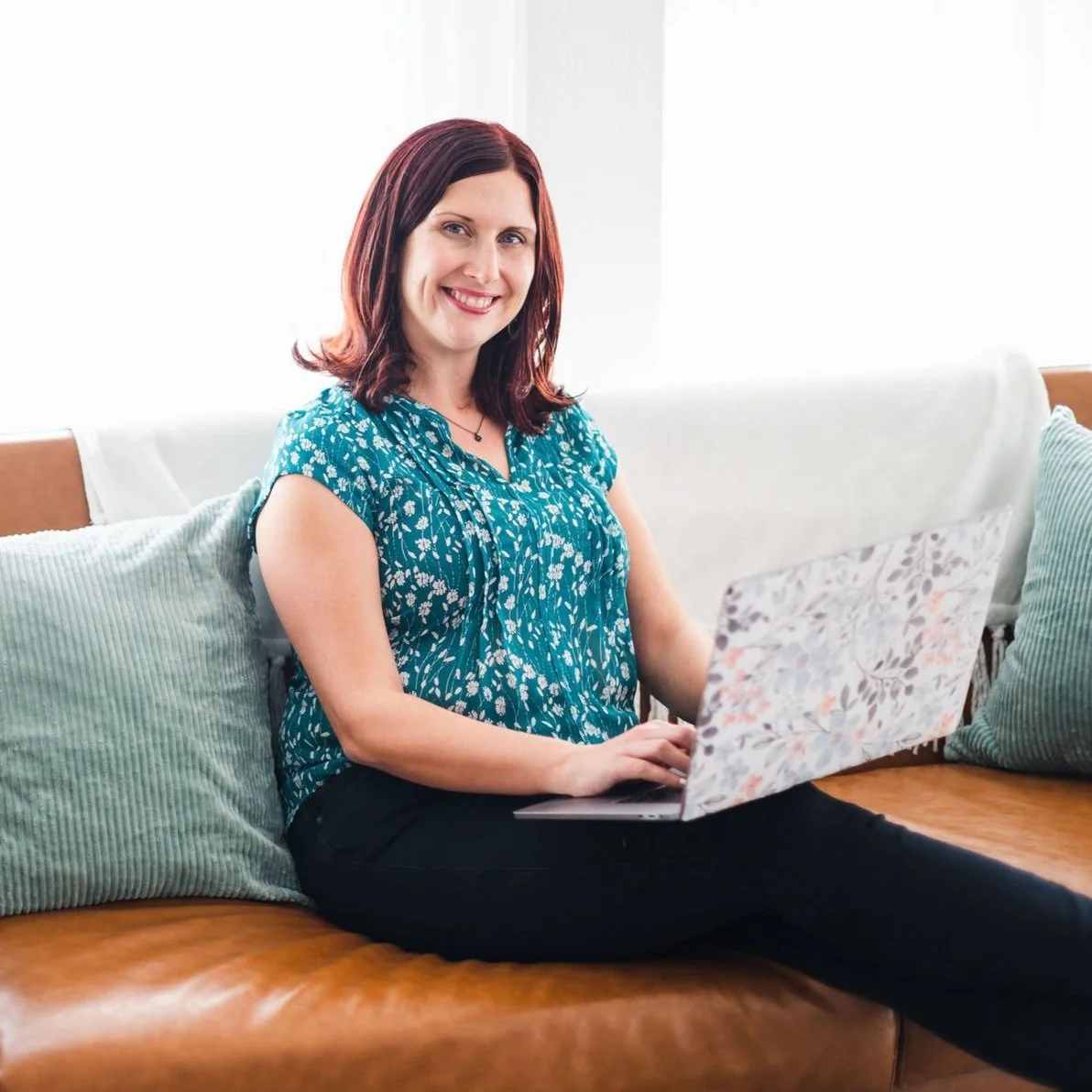 A woman with red hair and a floral blue top sitting on a leather couch with pillows, using a laptop, smiling at the camera.