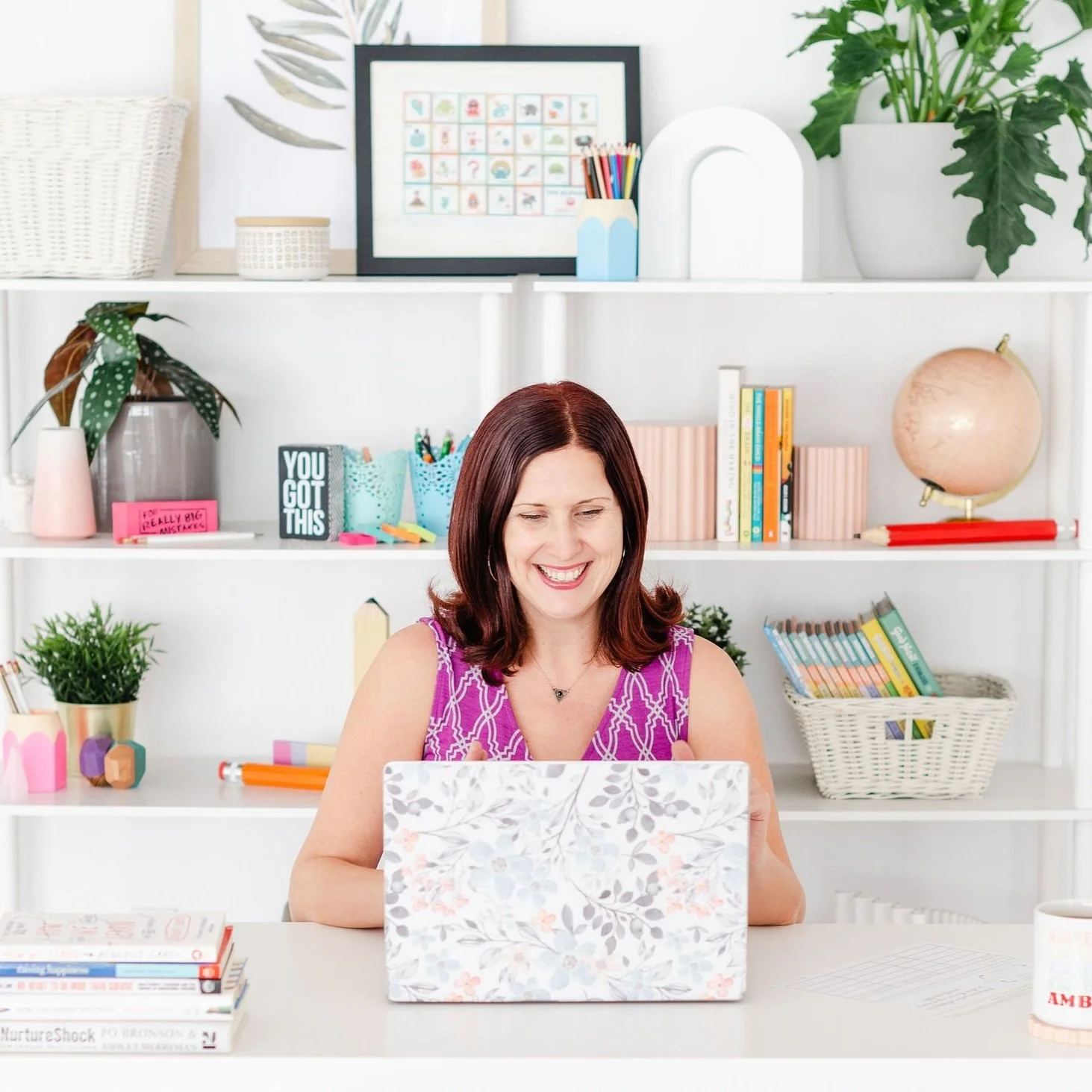 A woman with shoulder-length red hair smiling at a laptop in a bright room with white shelves filled with books, plants, and decorative items in the background.