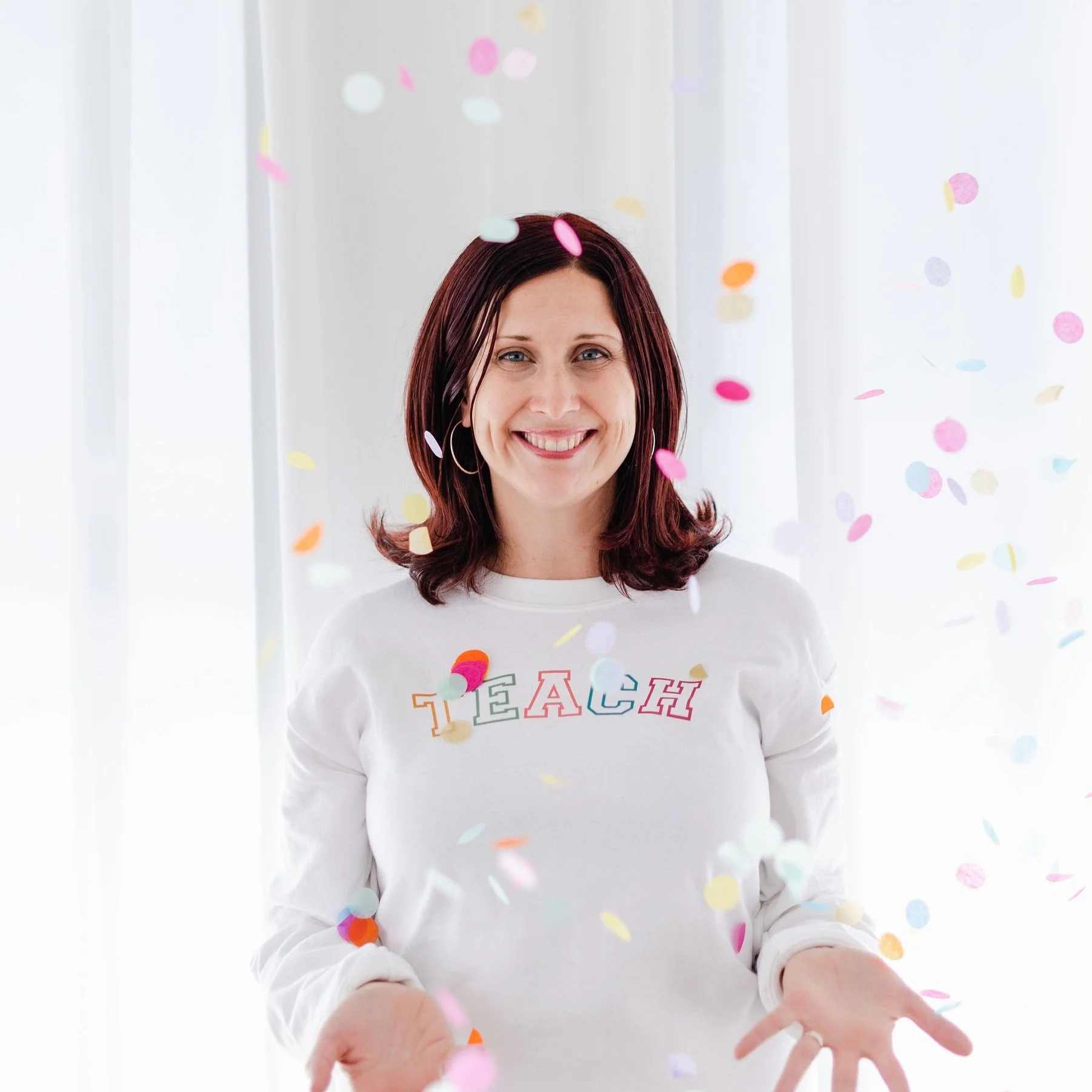 A woman with shoulder-length dark hair, smiling and wearing a white sweatshirt with the word 'TEACH' in colorful letters, is surrounded by falling colorful confetti in front of a bright, white background.