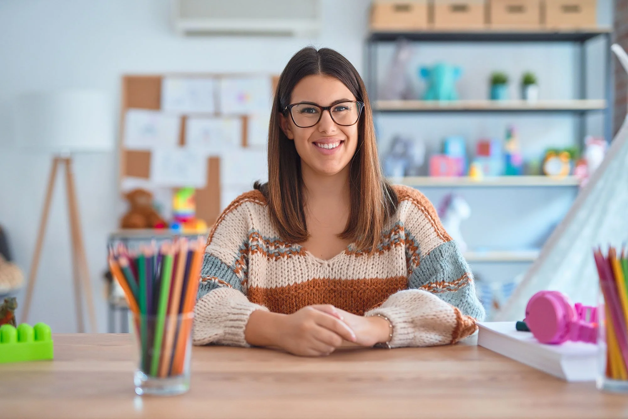 Smiling woman with glasses sitting at a desk in a creative space, surrounded by colorful art supplies and decorations.