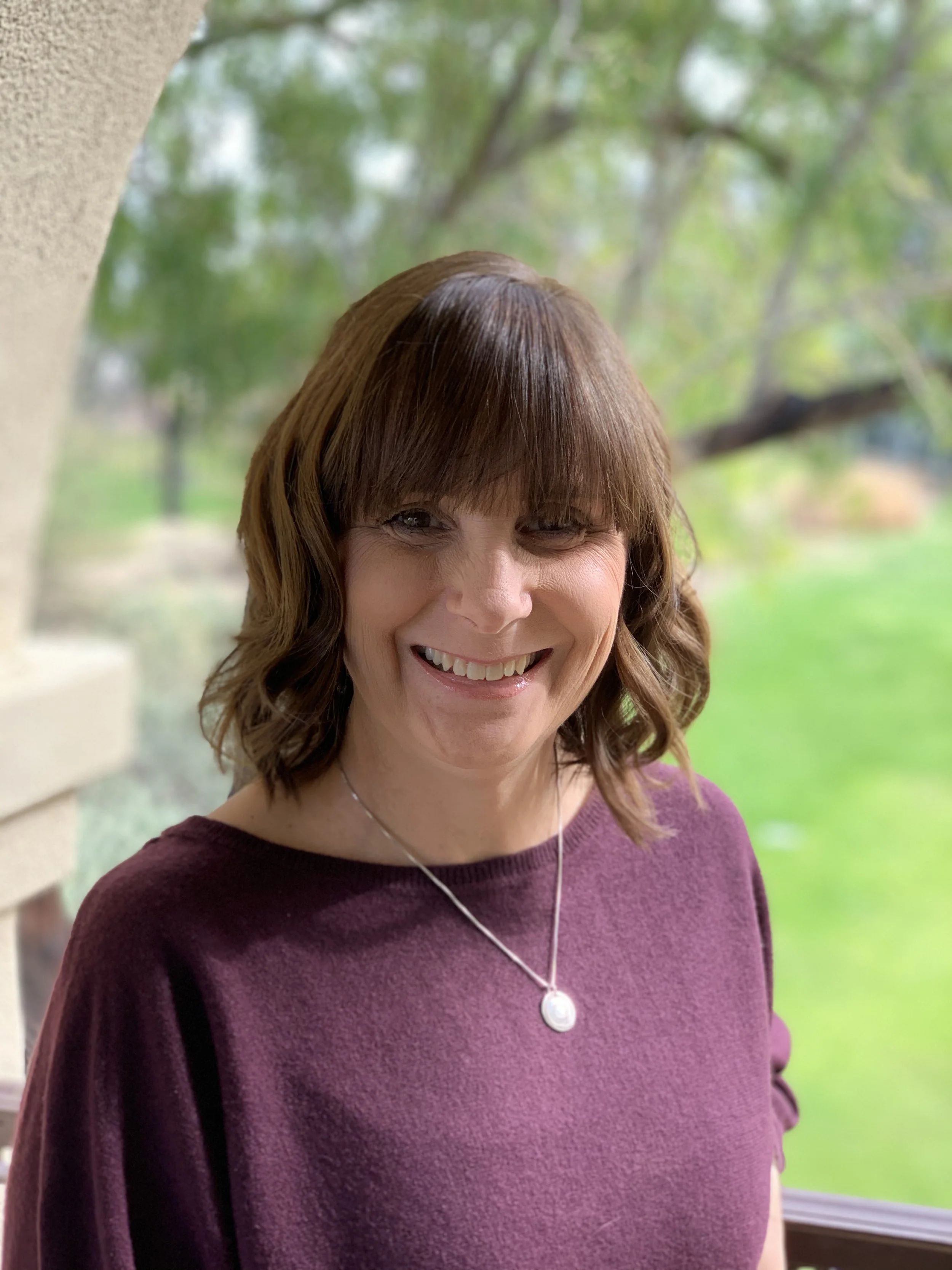 A woman with shoulder-length brown hair and bangs, smiling, standing outdoors near a window with a blurred green background of trees and grass, wearing a purple top and a silver necklace with a pendant.
