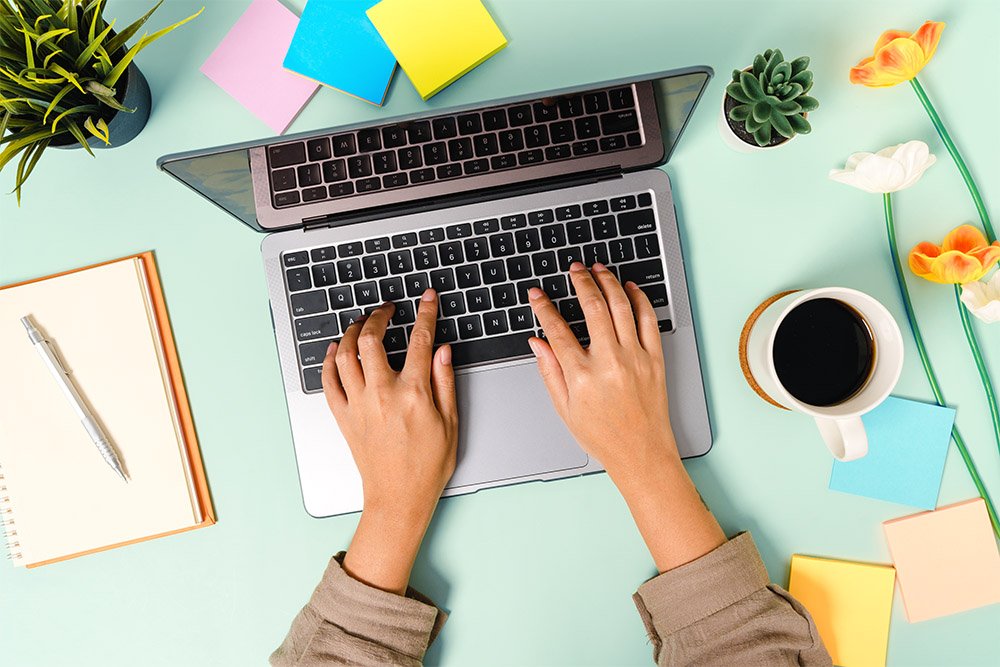 Person typing on a laptop keyboard surrounded by colorful sticky notes, flowers, a coffee cup, a notebook with a pen, and small potted plants on a light green desk.