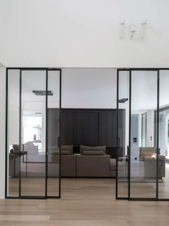 Modern living room visible through black-framed glass doors, with a gray sofa and wooden flooring.