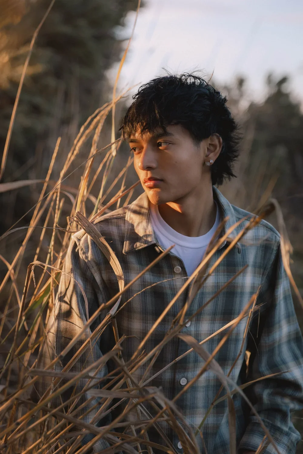A young man in a plaid shirt with a backpack, looking to the side amidst dry grass during sunset.