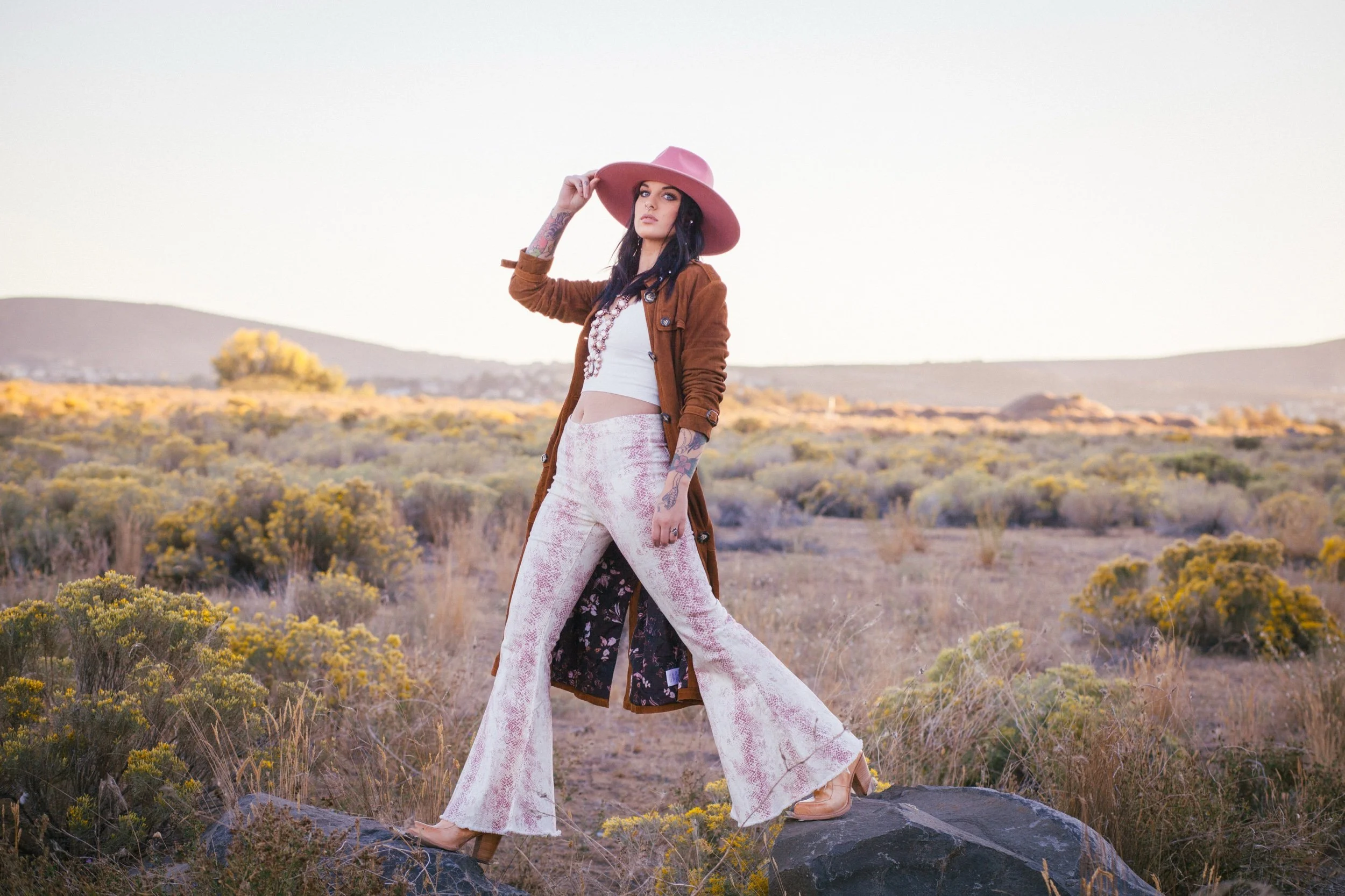 A woman with tattoos, wearing a wide pink hat, brown coat, white crop top, and pink patterned flared pants, standing on a rock in a desert landscape with yellow bushes and mountains in the background.