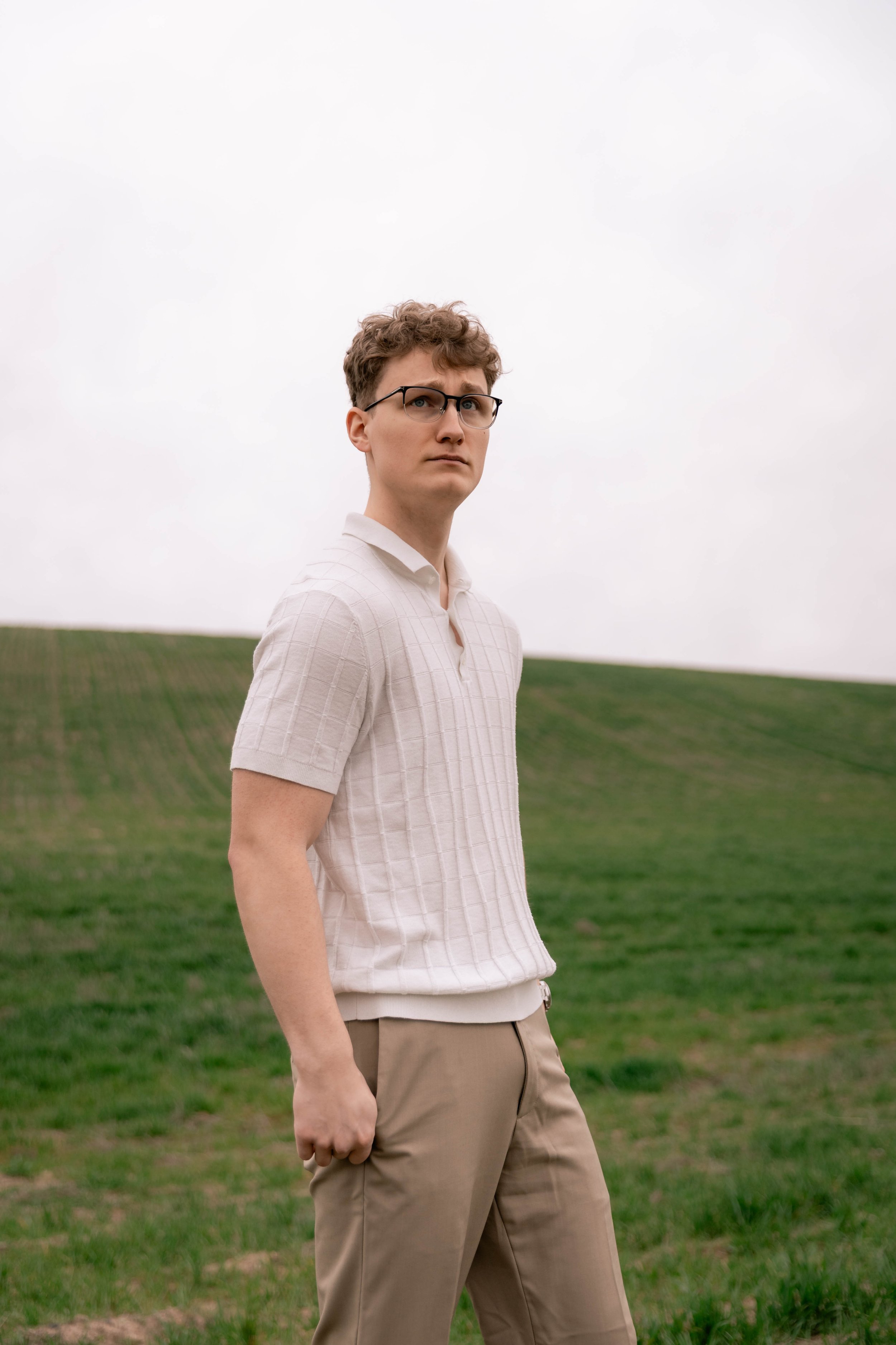 A young man stands in a grassy field on an overcast day, wearing glasses, a light-colored short-sleeve polo shirt, and beige pants, looking to the side with a serious expression.
