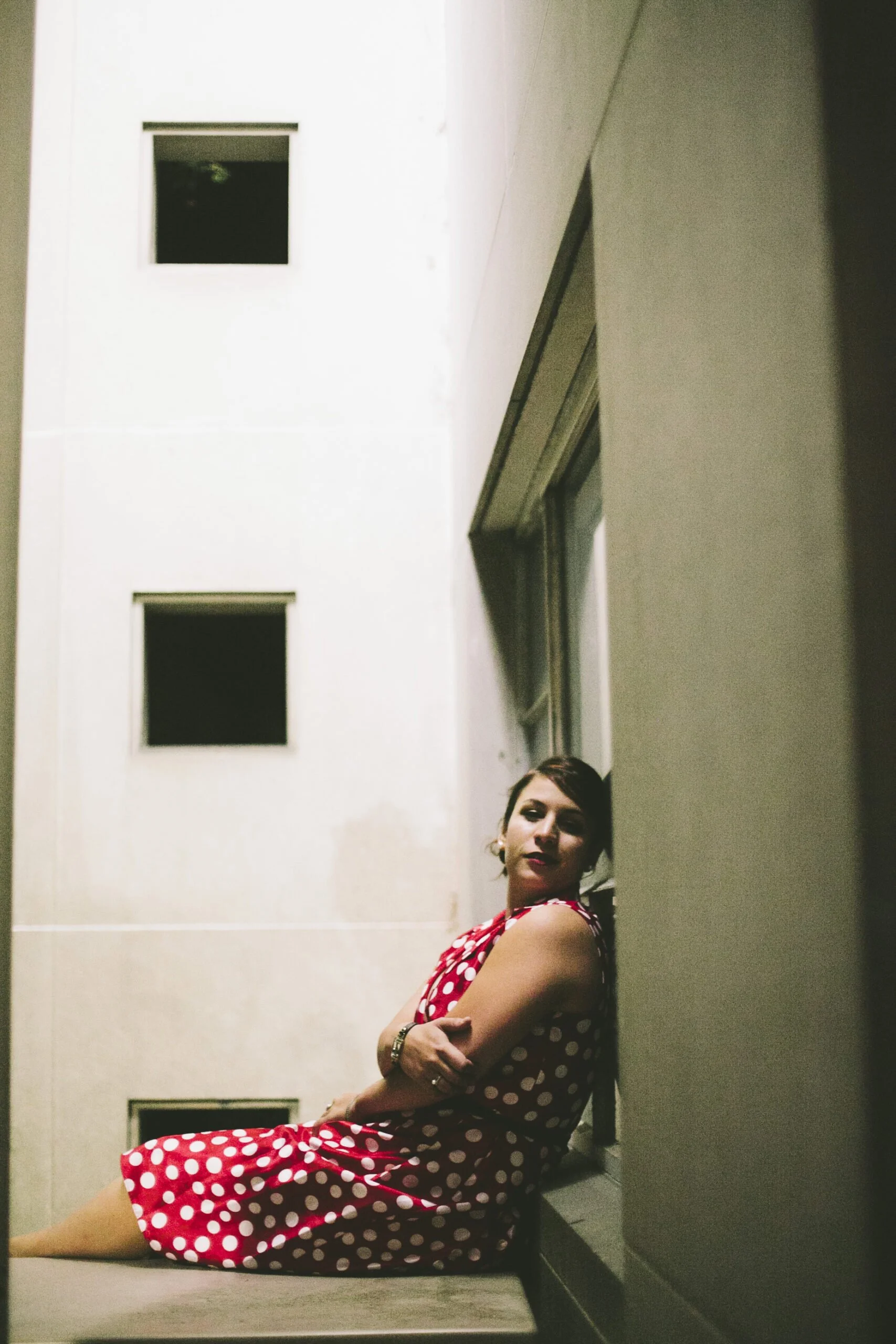 A woman in a red polka dot dress sitting on a ledge outside a building at night, with her arms crossed and looking at the camera.