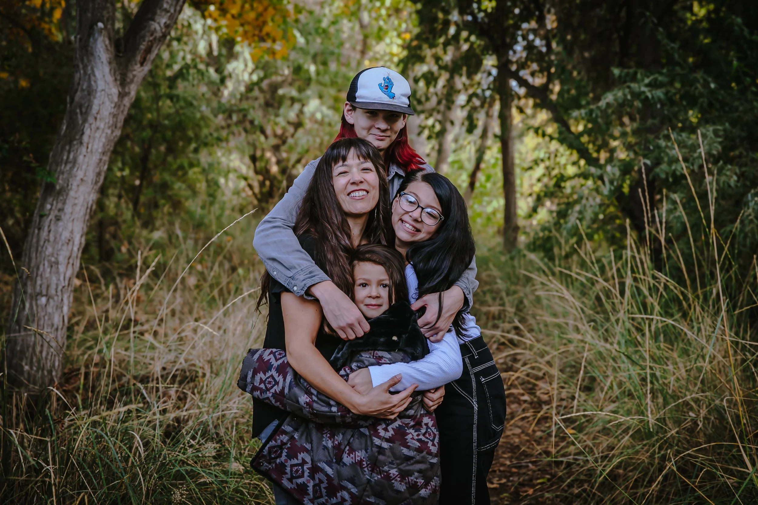 Family of four, including two young girls, an adult woman, and a teenage girl, hugging outdoors in a wooded area.