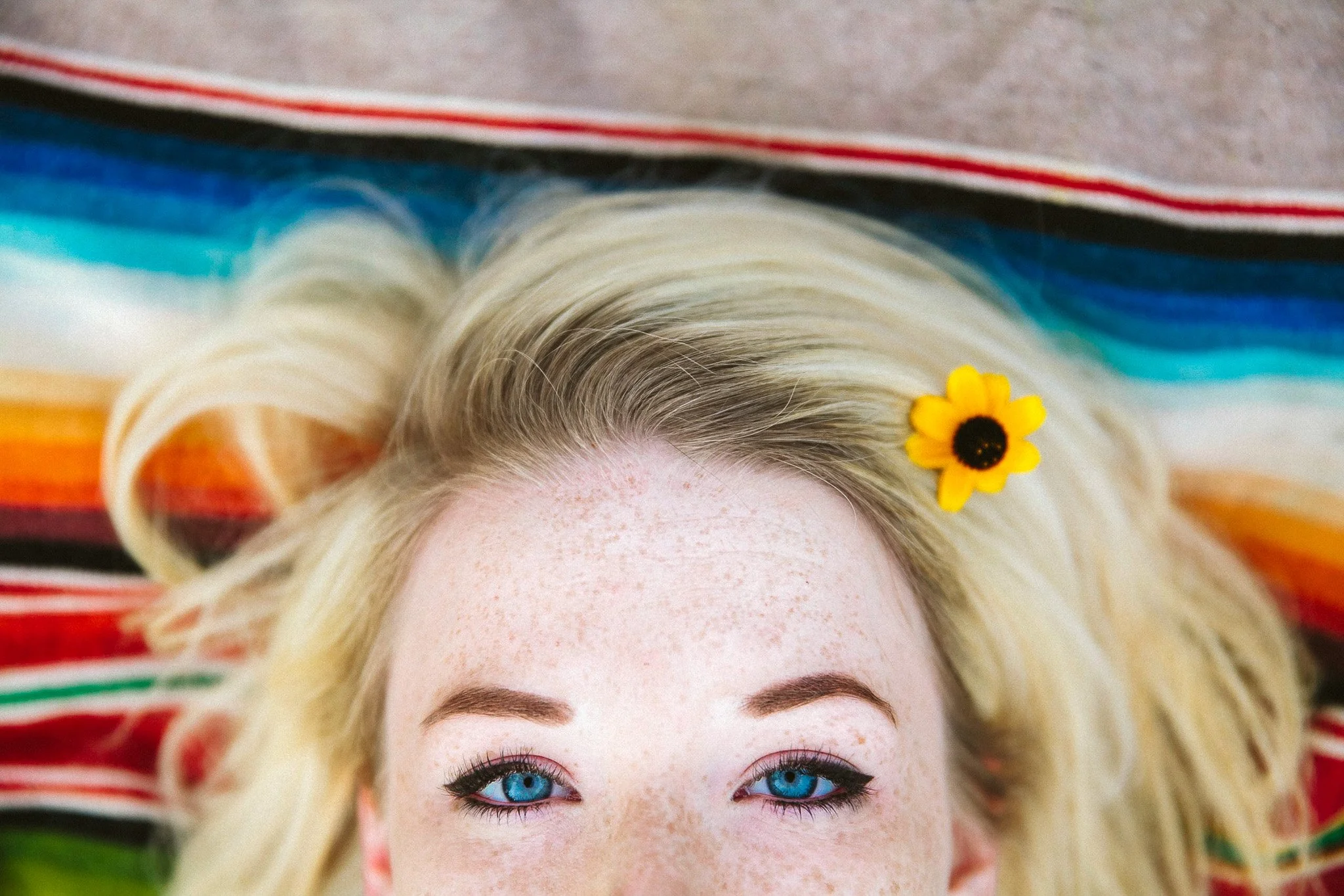A woman lying on a multicolored striped blanket with her blonde hair spread out around her, a yellow flower near her temple, and bright blue eyes.