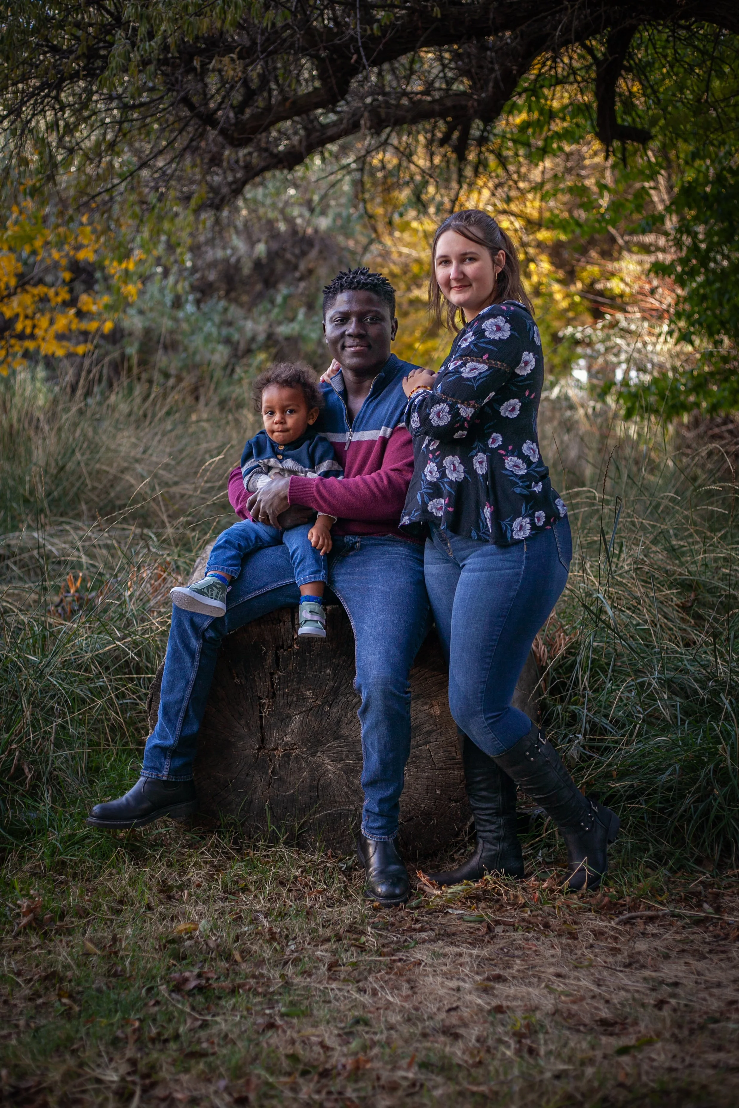 A family of three sitting on a tree stump outdoors surrounded by trees and foliage during autumn, with a man holding a young child on his lap and a woman standing next to them.