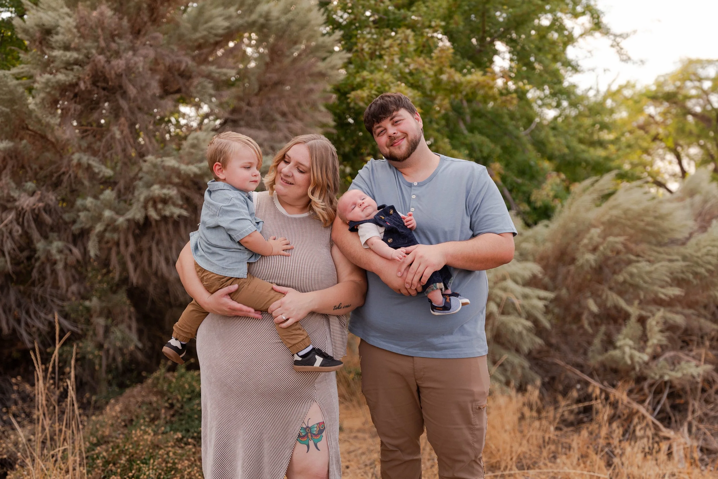 Family of four, including a woman, a man, a young boy, and a baby, posing outdoors in front of trees during sunset.