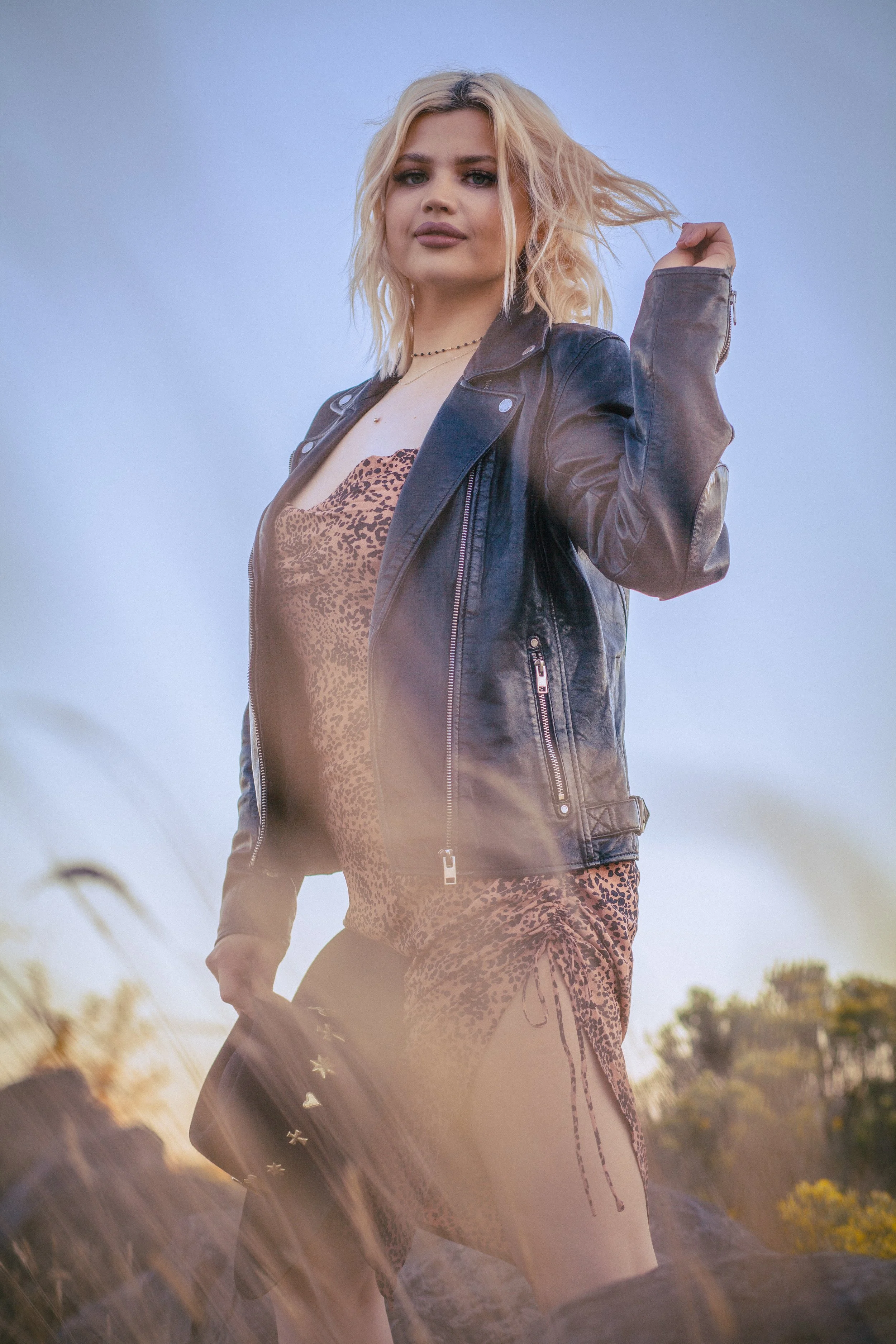 A young woman with blonde hair wearing a black leather jacket and a leopard print dress outdoors during sunset, with tall grass and trees in the background.