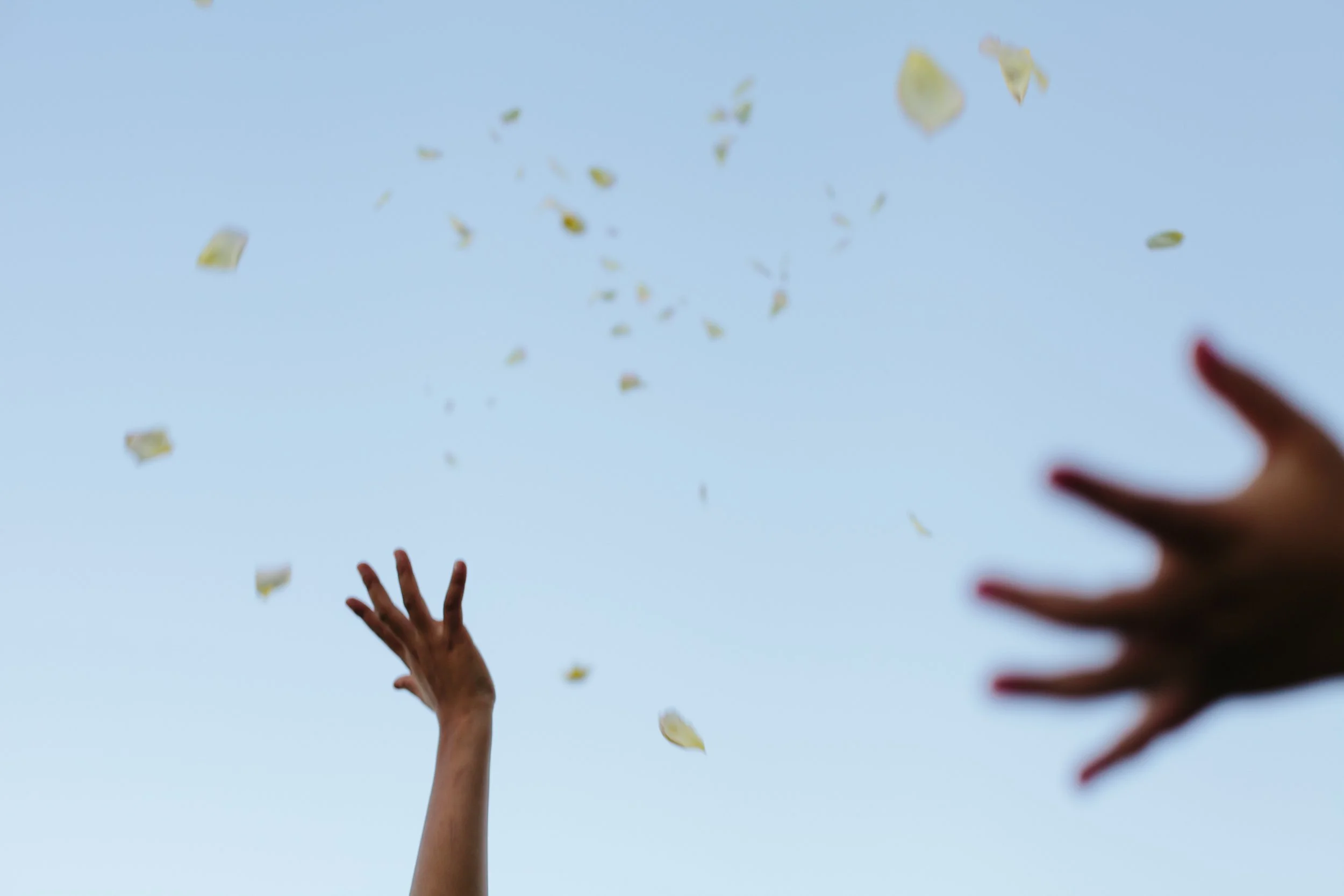 Two hands reaching up toward falling leaves against a clear blue sky.
