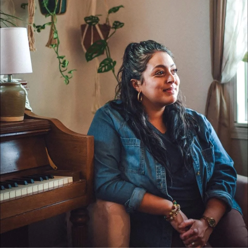 A woman with dark hair and hoop earrings sitting on a beige armchair, in a room with a piano, potted plants, and a window with curtains.