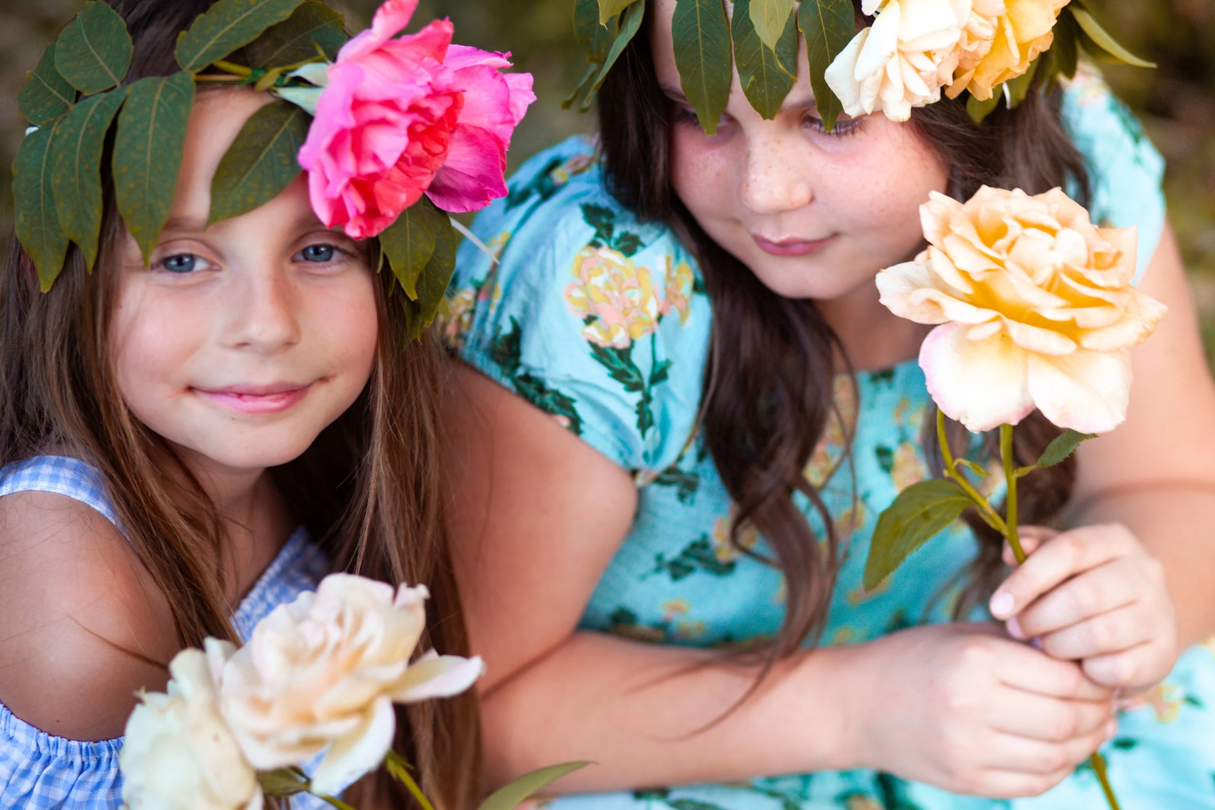 Two young girls with floral crowns made of large roses and leaves, holding roses, outdoors.