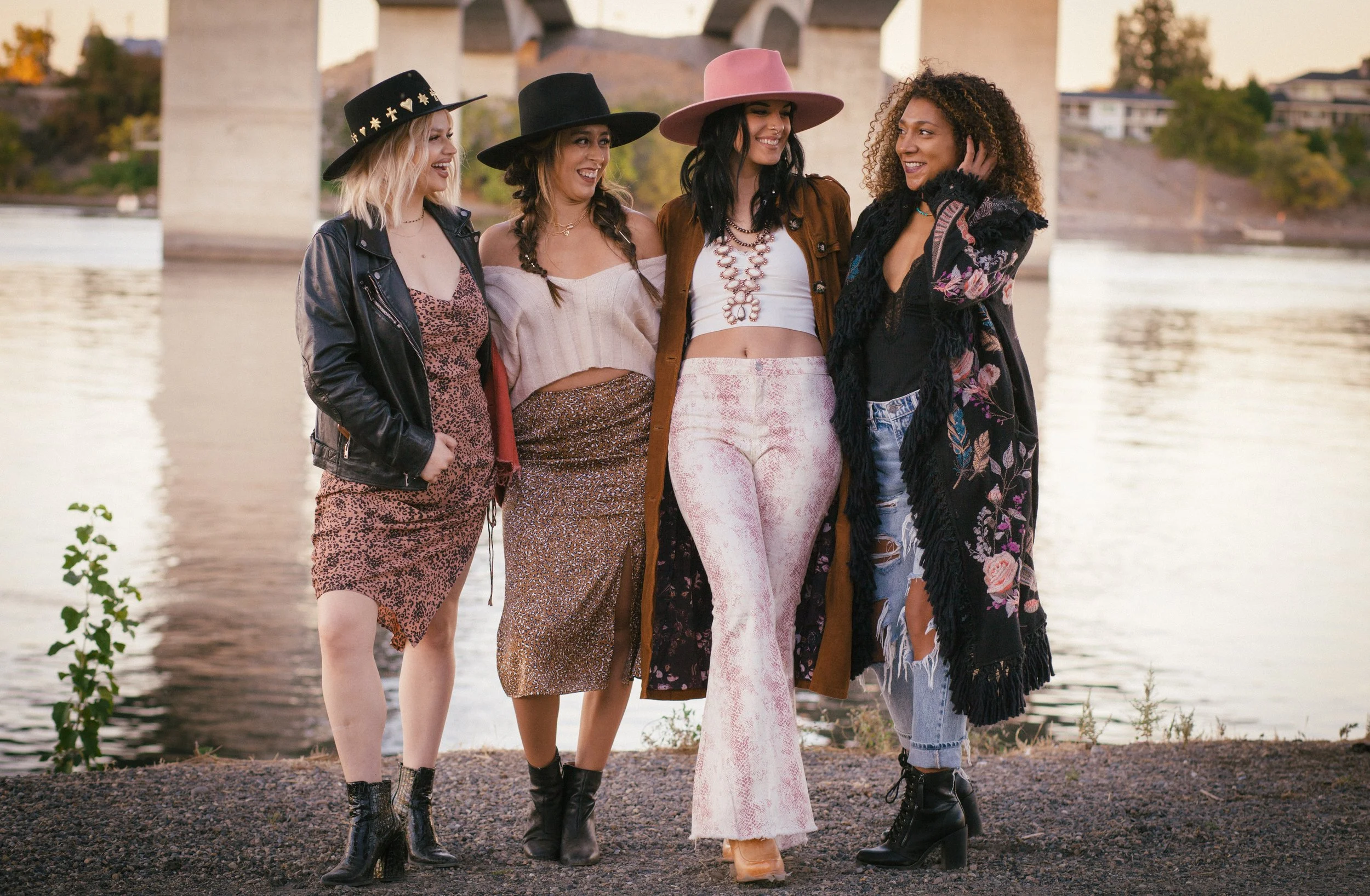 Five women walking together near a river, smiling and dressed in fashionable clothing and wide-brimmed hats.