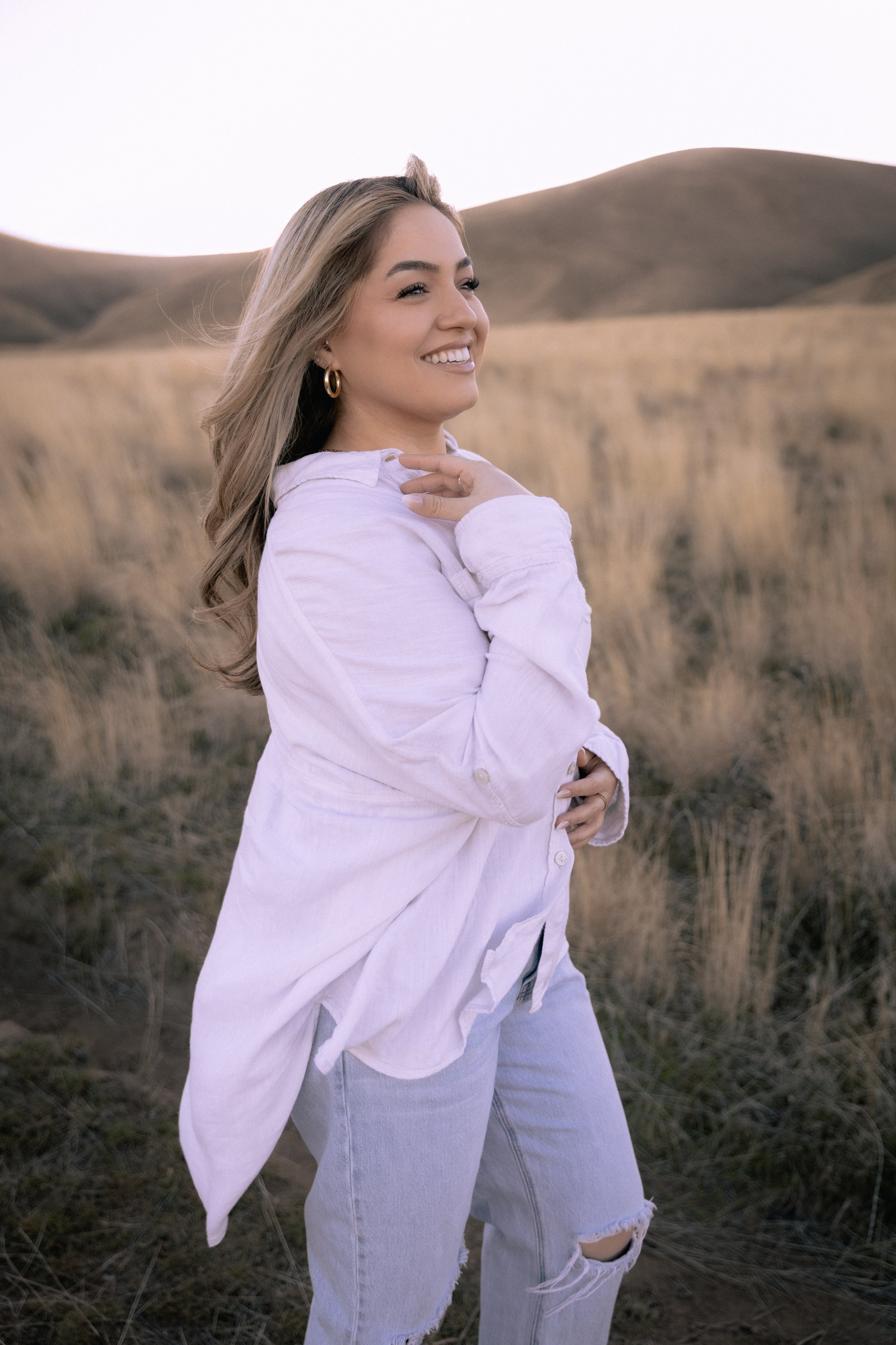 A smiling young woman with long hair wearing a white shirt and jeans standing in a grassy outdoor field with rolling hills in the background.