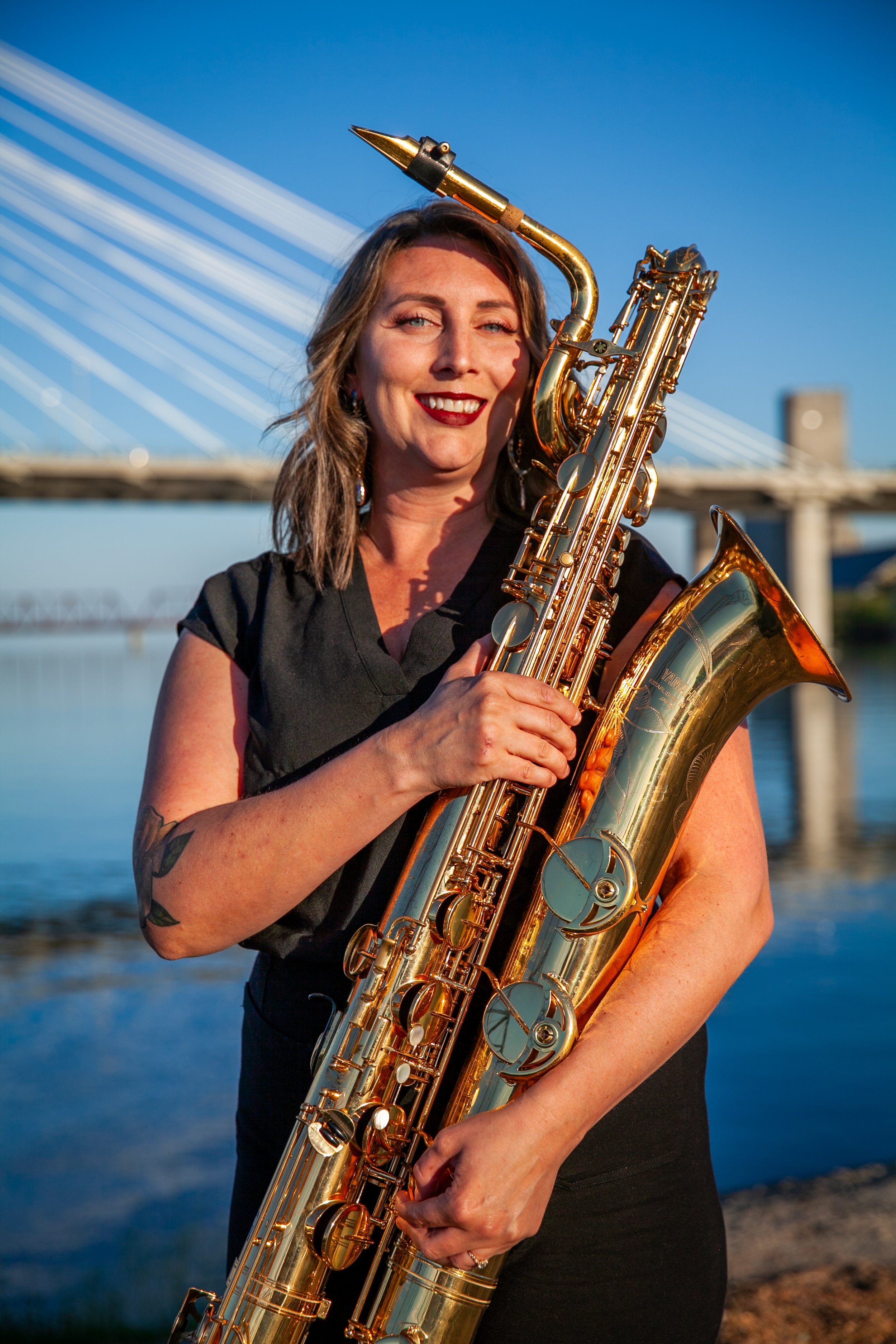 A woman holding a saxophone outdoors near a river with a bridge in the background during sunset, smiling at the camera.
