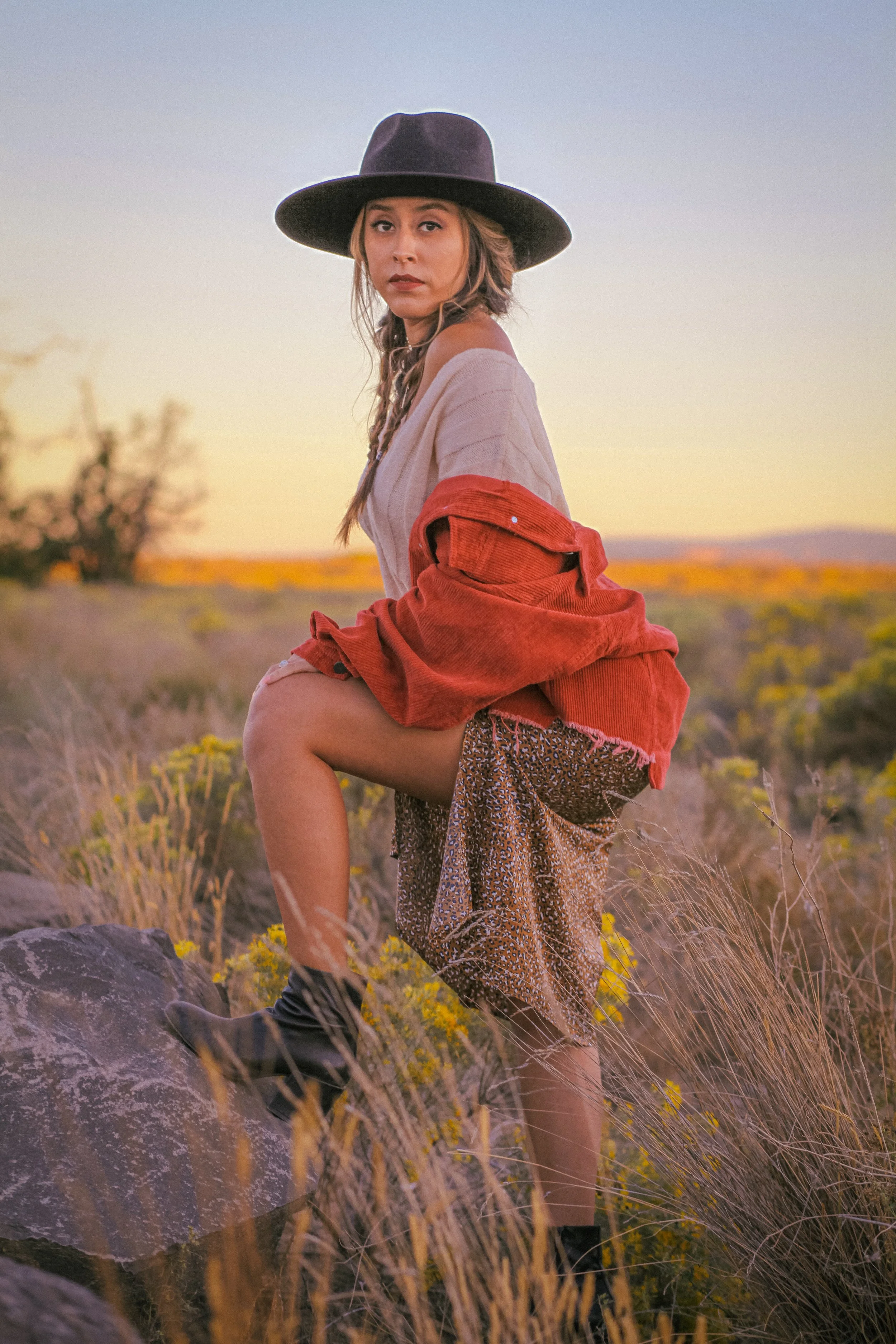 A woman wearing a wide-brimmed hat, tan sweater, a red jacket, patterned skirt, and combat boots stands outdoors in a field with rocks and yellow wildflowers, during sunset with a distant horizon.